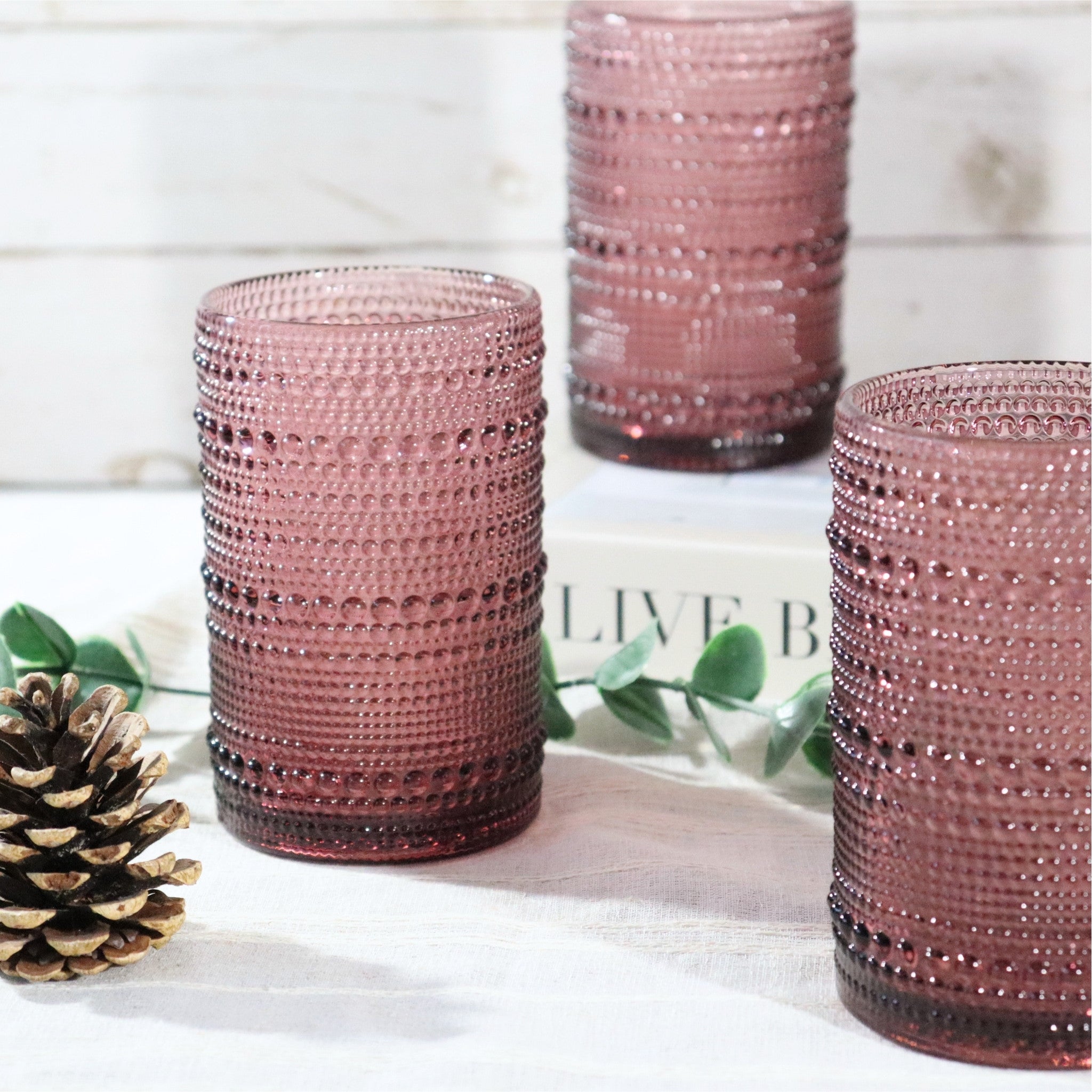 Three purple textured glass tumblers on a white surface with greenery and a pine cone.