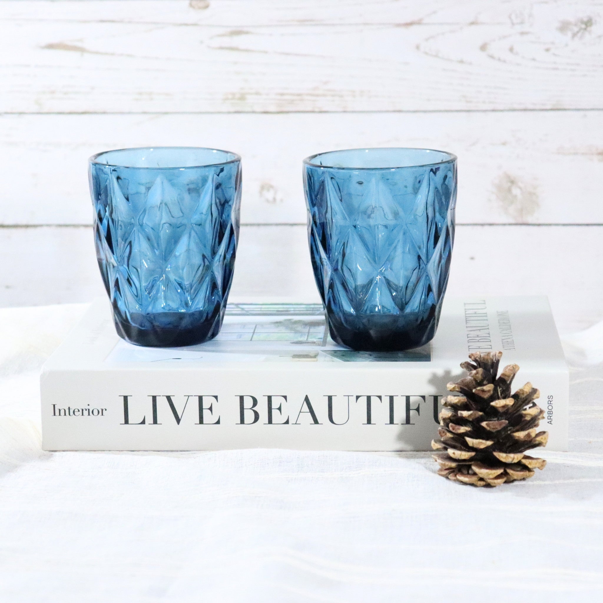 Two blue textured glass tumblers on a book with a pine cone on a white surface.