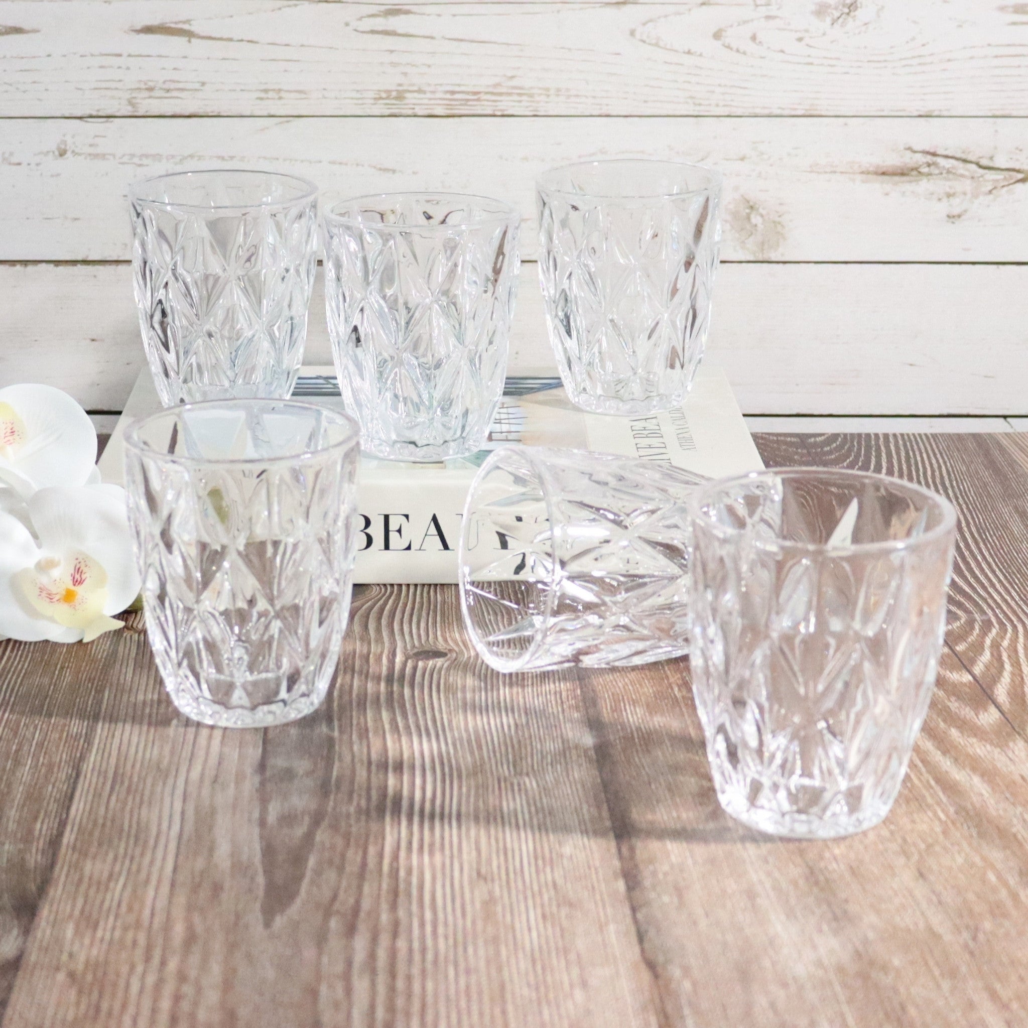 Set of six clear glass tumblers on a wooden surface with a blurred 'BEA' book in the background.