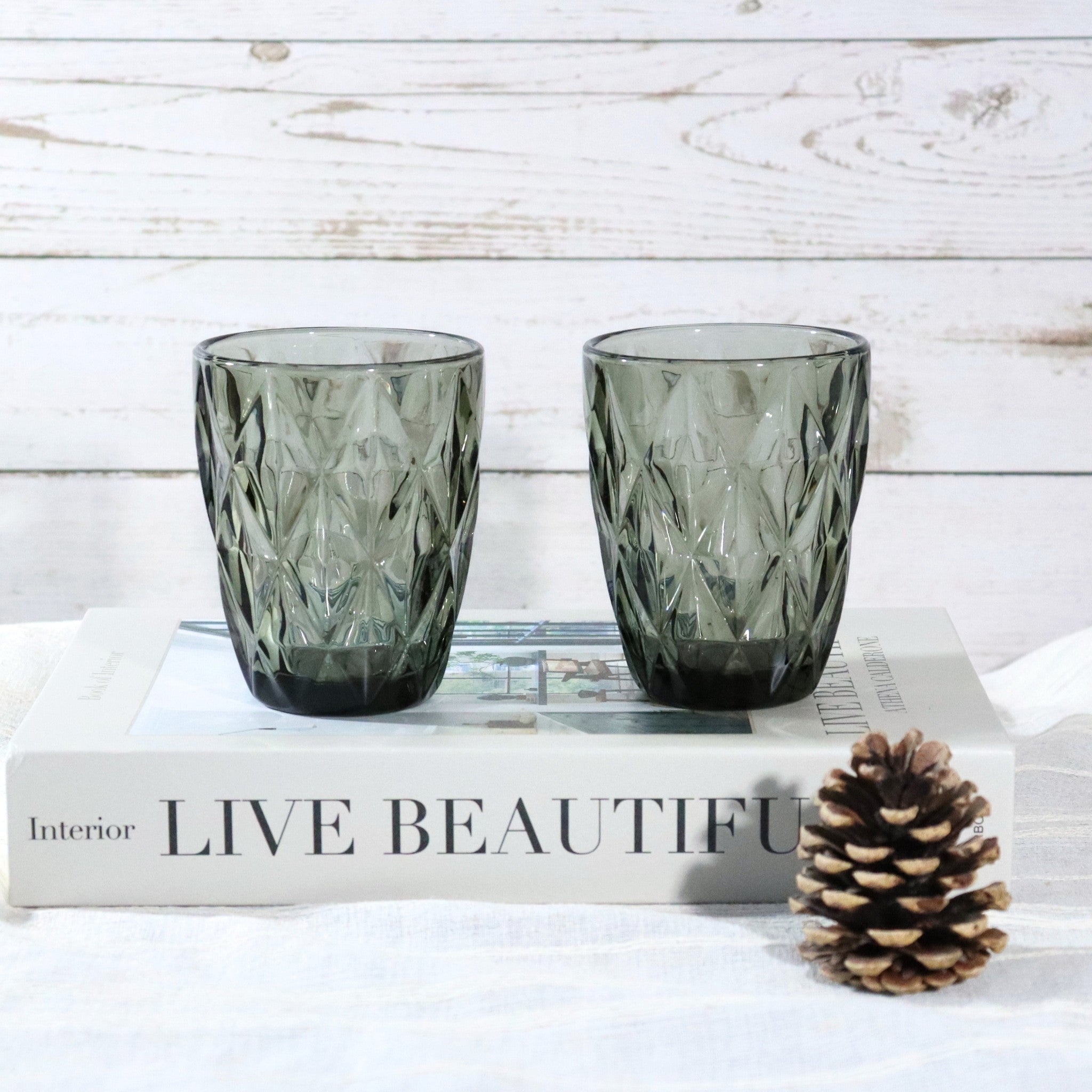 Two green textured glasses on a book with a pine cone on a white surface.