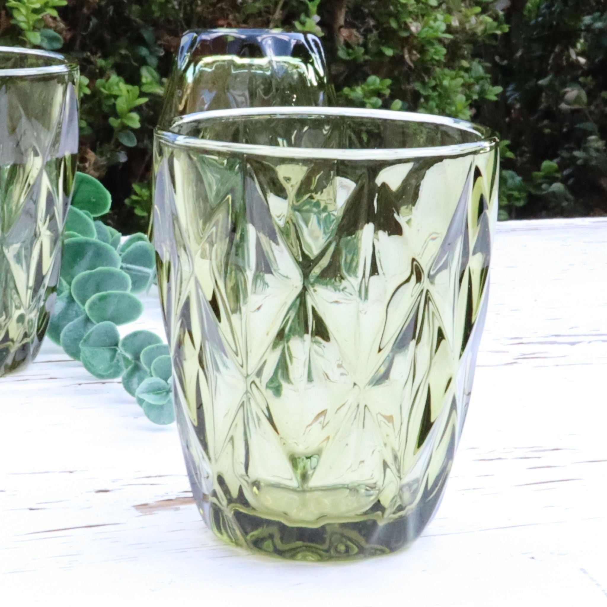 Green glass tumblers with diamond pattern on a white surface with greenery in the background