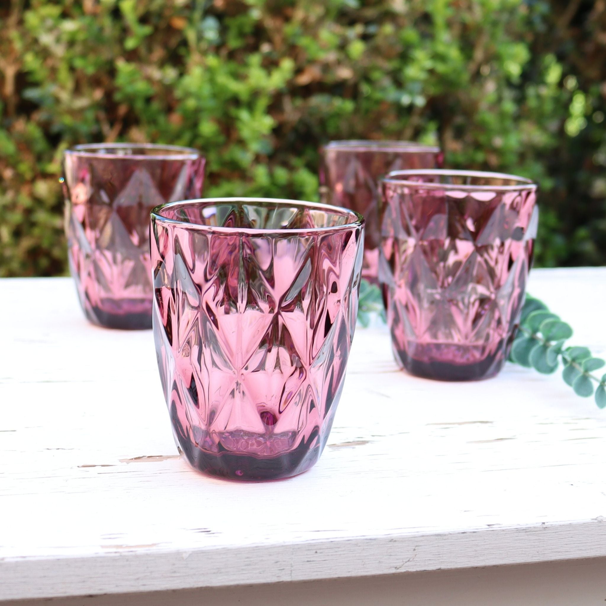 Set of four purple glass tumblers on a white surface with greenery in the background