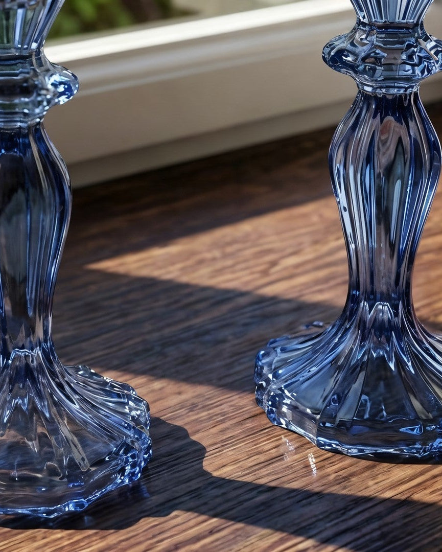 Two blue glass candlestick holders with a fluted, faceted design, displayed on a wooden windowsill in natural sunlight.