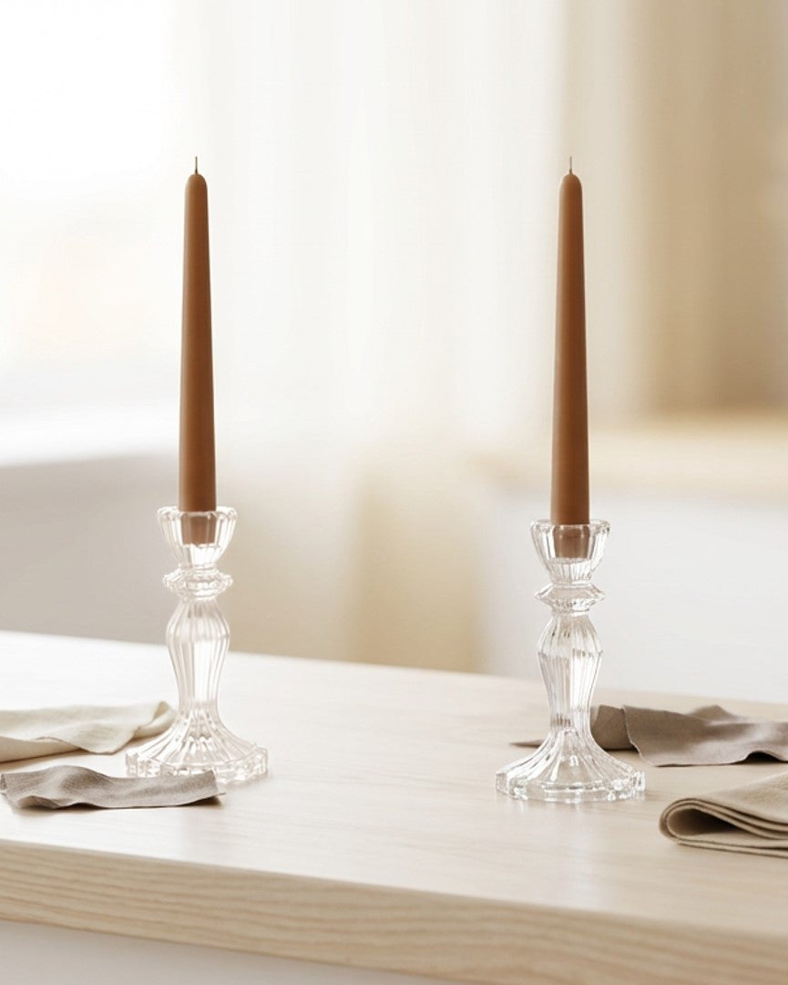 Two candlesticks with brown candles on a white surface near a window.