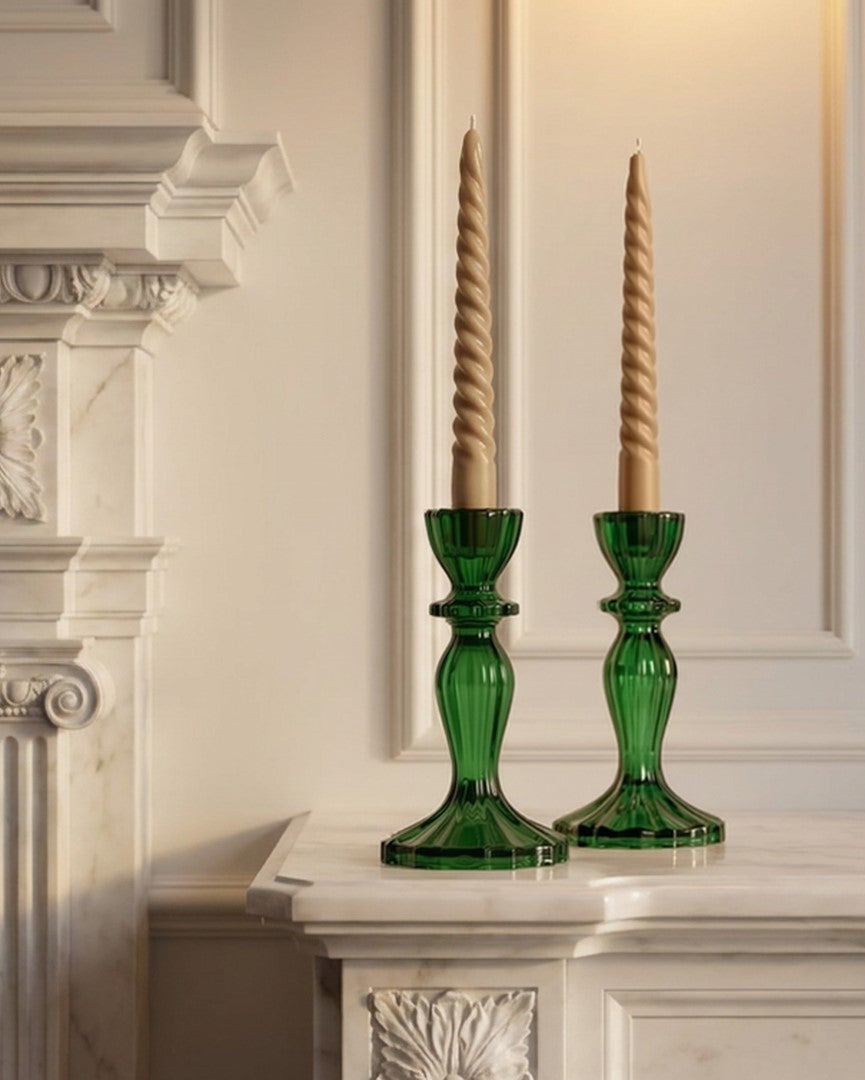 Two green glass candlestick holders with twisted beige taper candles, displayed on a white marble mantelpiece in front of a classic white fireplace.