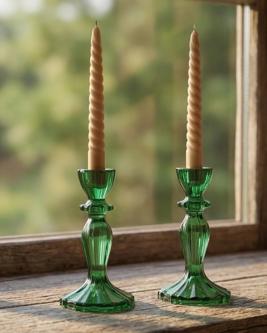 Two green glass candlestick holders with twisted cream taper candles, displayed on a rustic wooden windowsill with a blurred outdoor forest background.