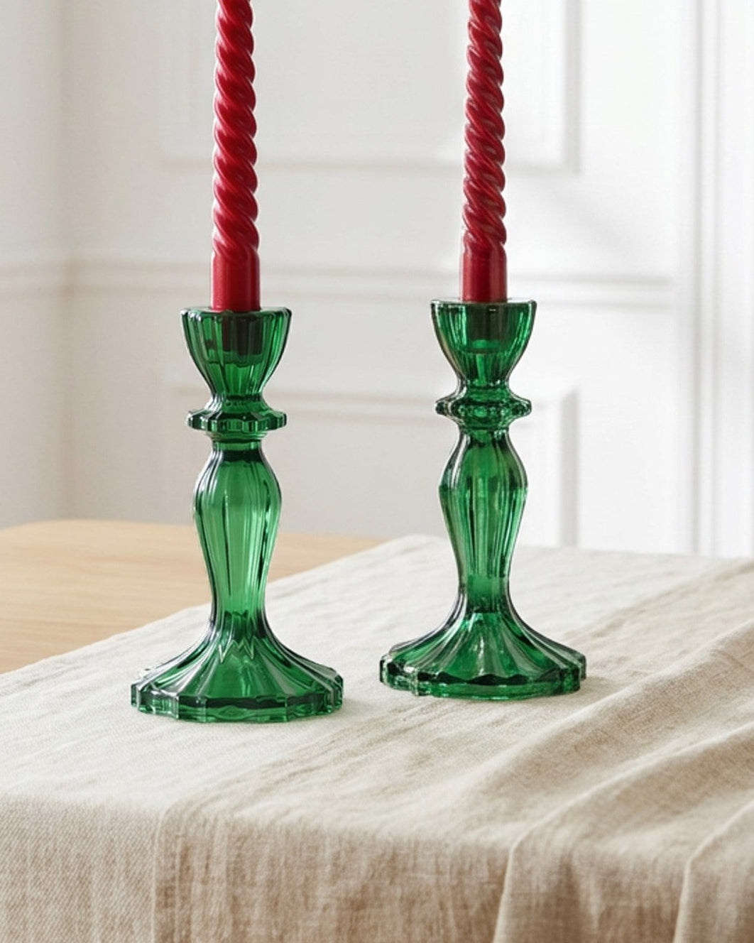 Two green glass candlesticks with red candles on a wooden surface with white flowers.