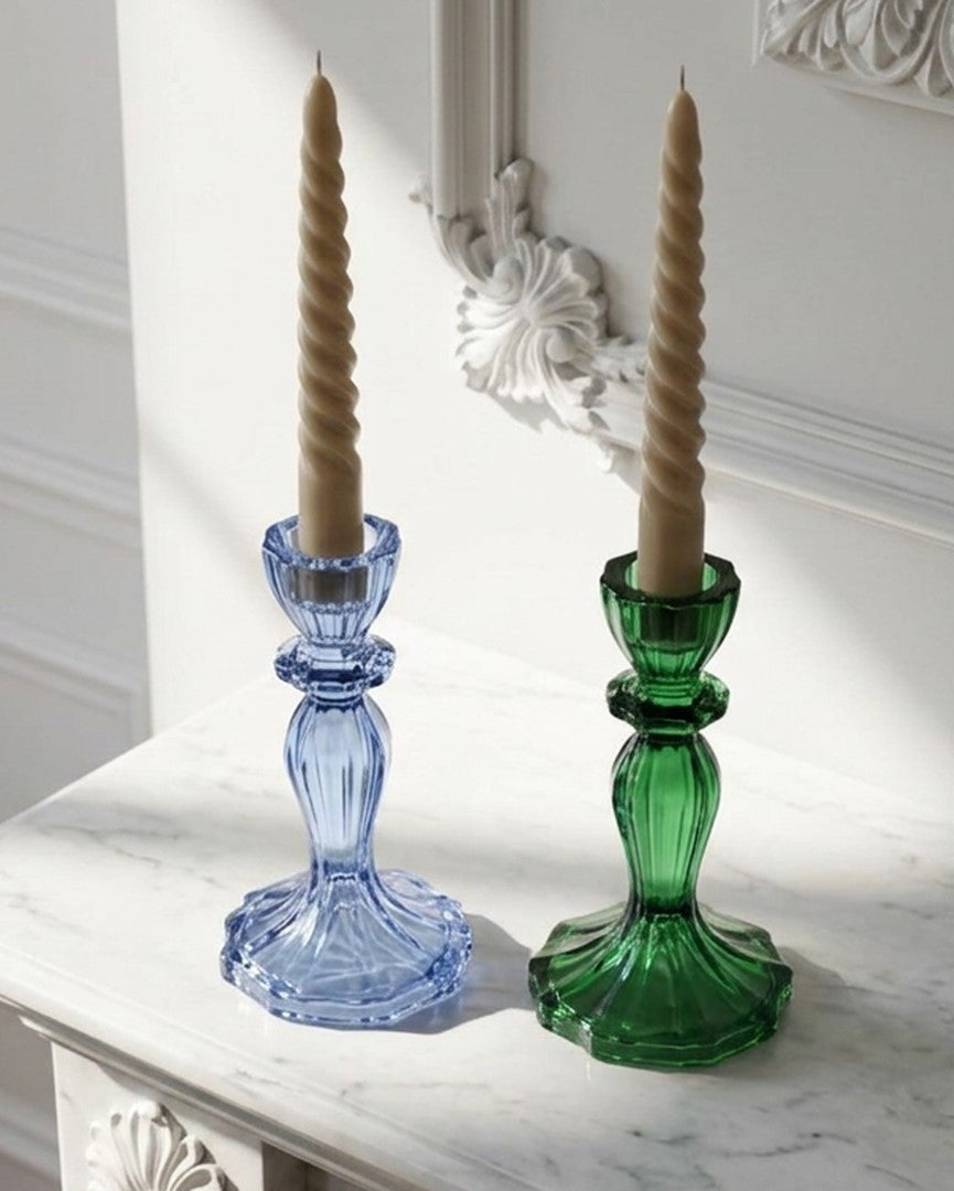 Two glass candlestick holders, one blue and one green, holding twisted beige taper candles, set on a white marble console table against a white paneled wall.