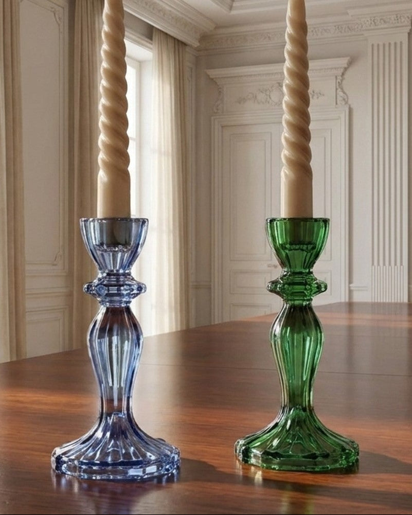 Two twisted taper candles in blue and green glass candlesticks on a polished wooden table in an elegant room with cream walls and tall windows.