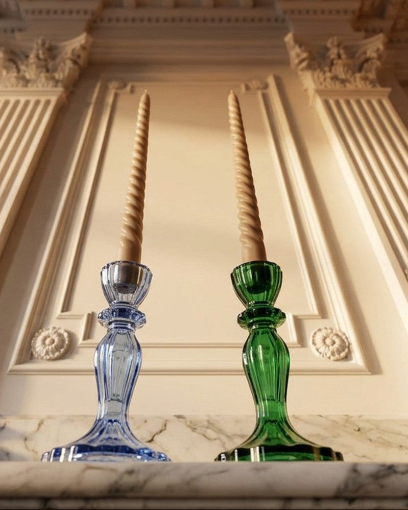 Two decorative glass candlesticks, one blue and one green, holding twisted taper candles, set against an ornate cream-colored classical wall with fluted pilasters.