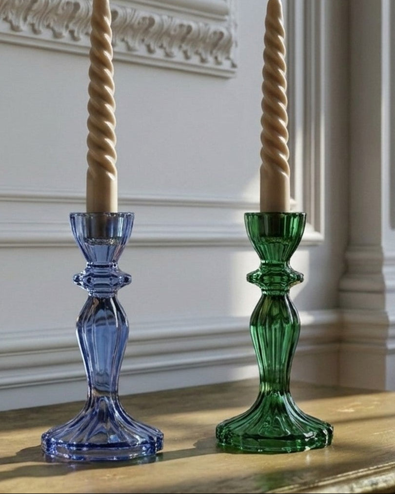 Two decorative glass candlesticks, one blue and one green, holding twisted cream taper candles, displayed on a wooden console table in a classical room with white walls and molding.