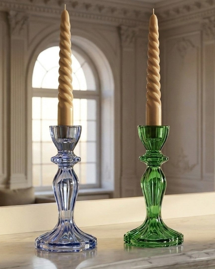 Two elegant twisted taper candlestick holders on a marble mantelpiece, one in translucent blue glass and one in translucent green glass, set against a classical interior backdrop with columns and arched windows.