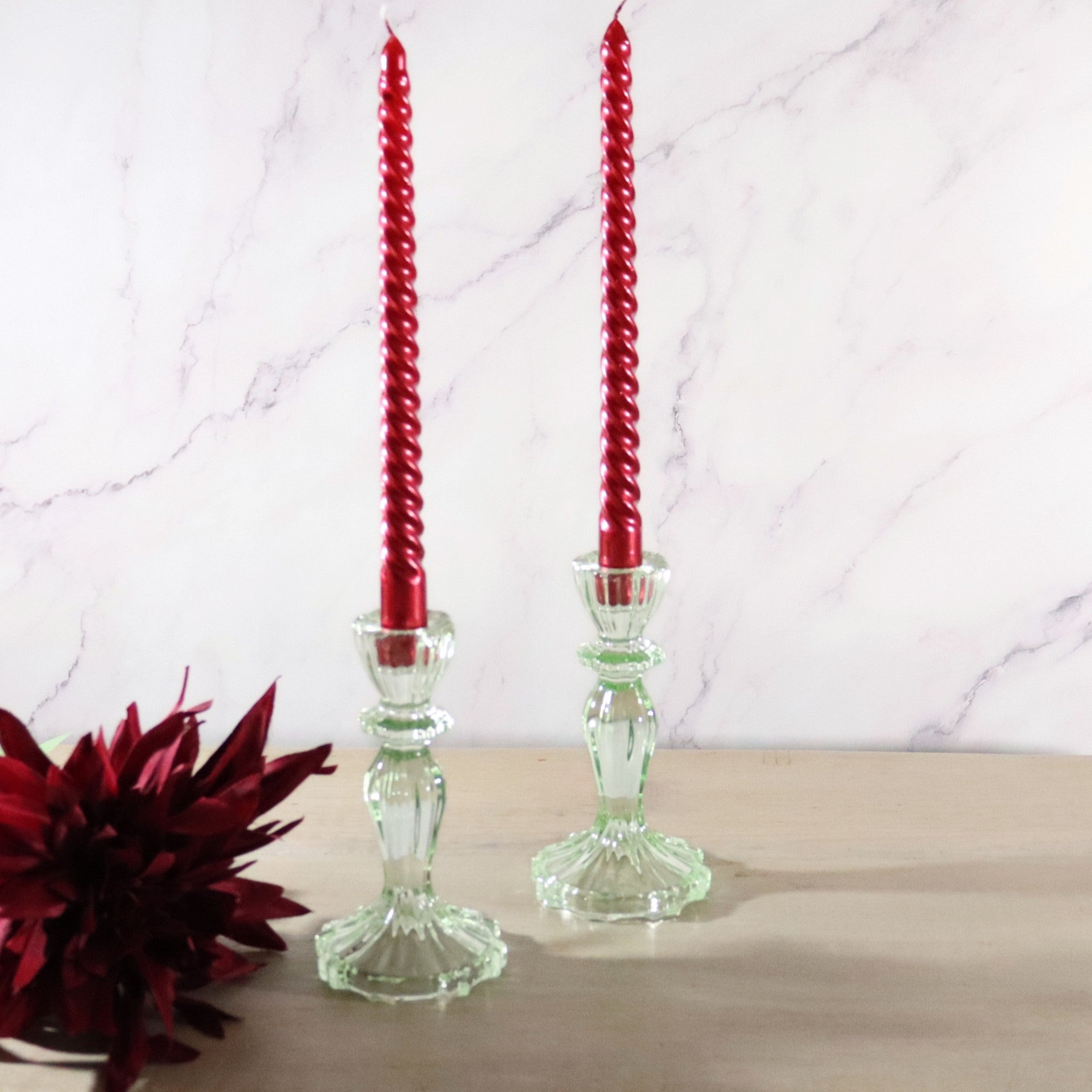 Two twisted candles in light green glass candle holder on a marble surface with a red flower.