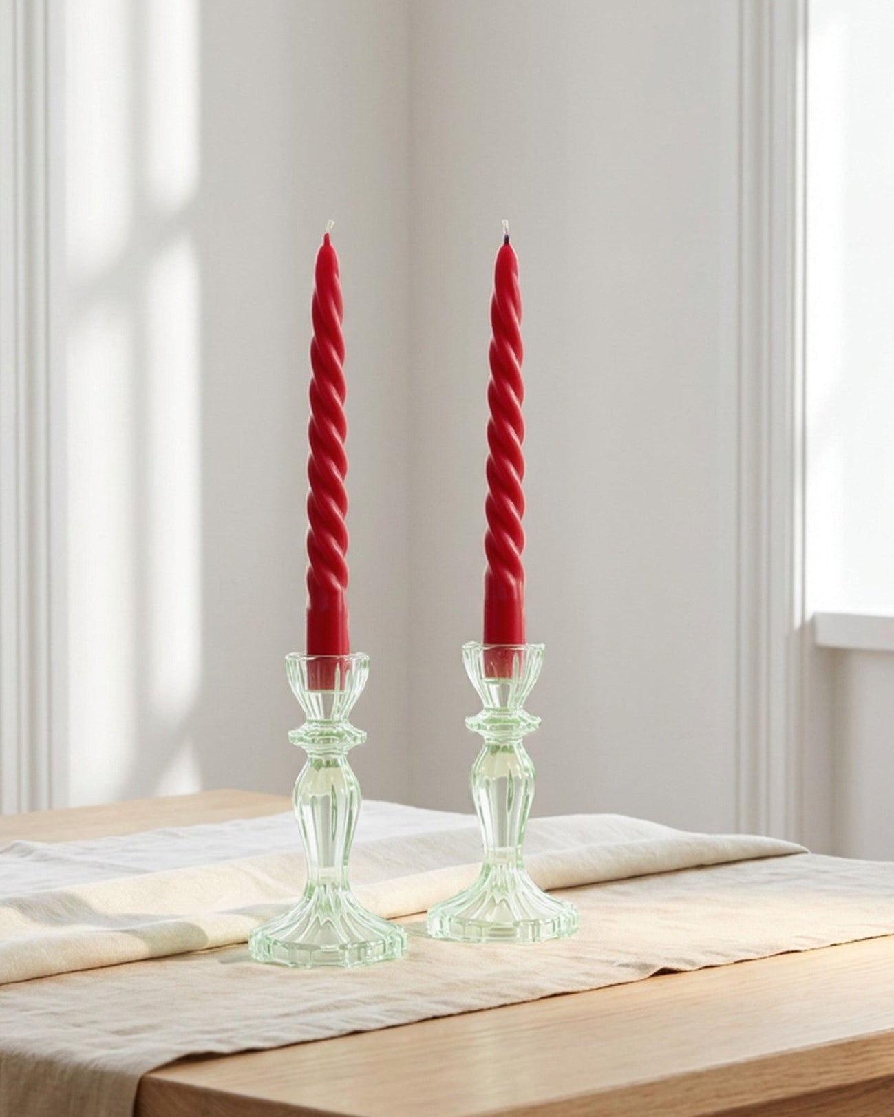 Two red candles in green candlesticks on a wooden table with a neutral tablecloth.