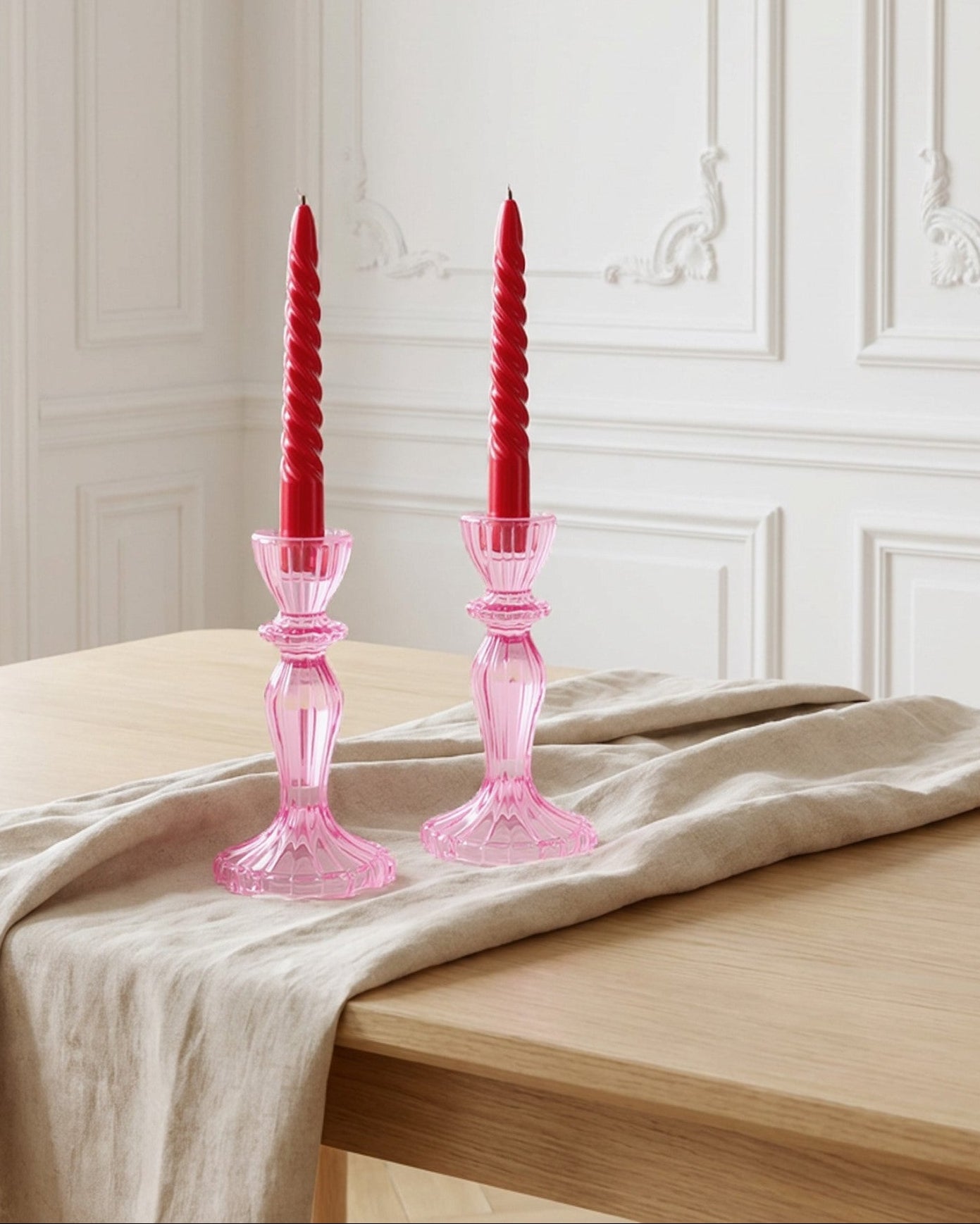 Pink glass candle holders with red candles on a wooden surface with white flowers.