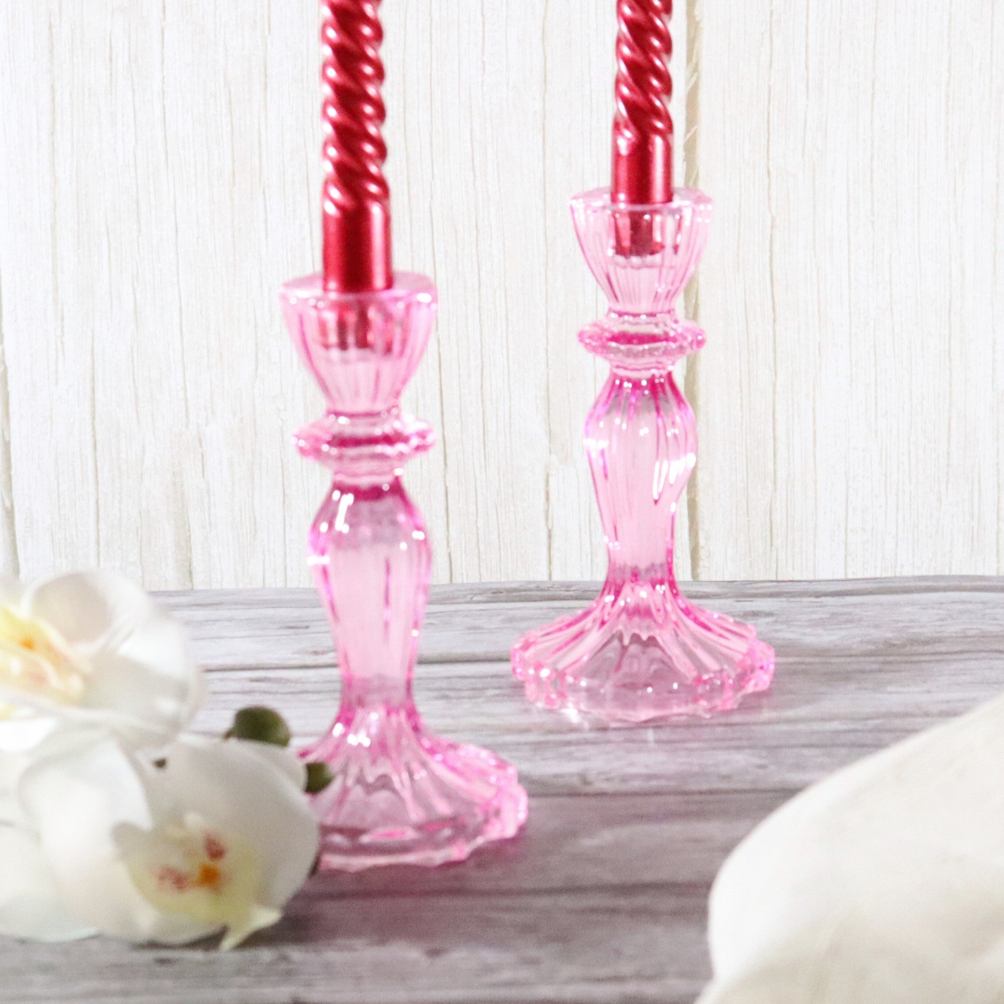Pink glass candle holders with red candles on a wooden surface with white flowers.