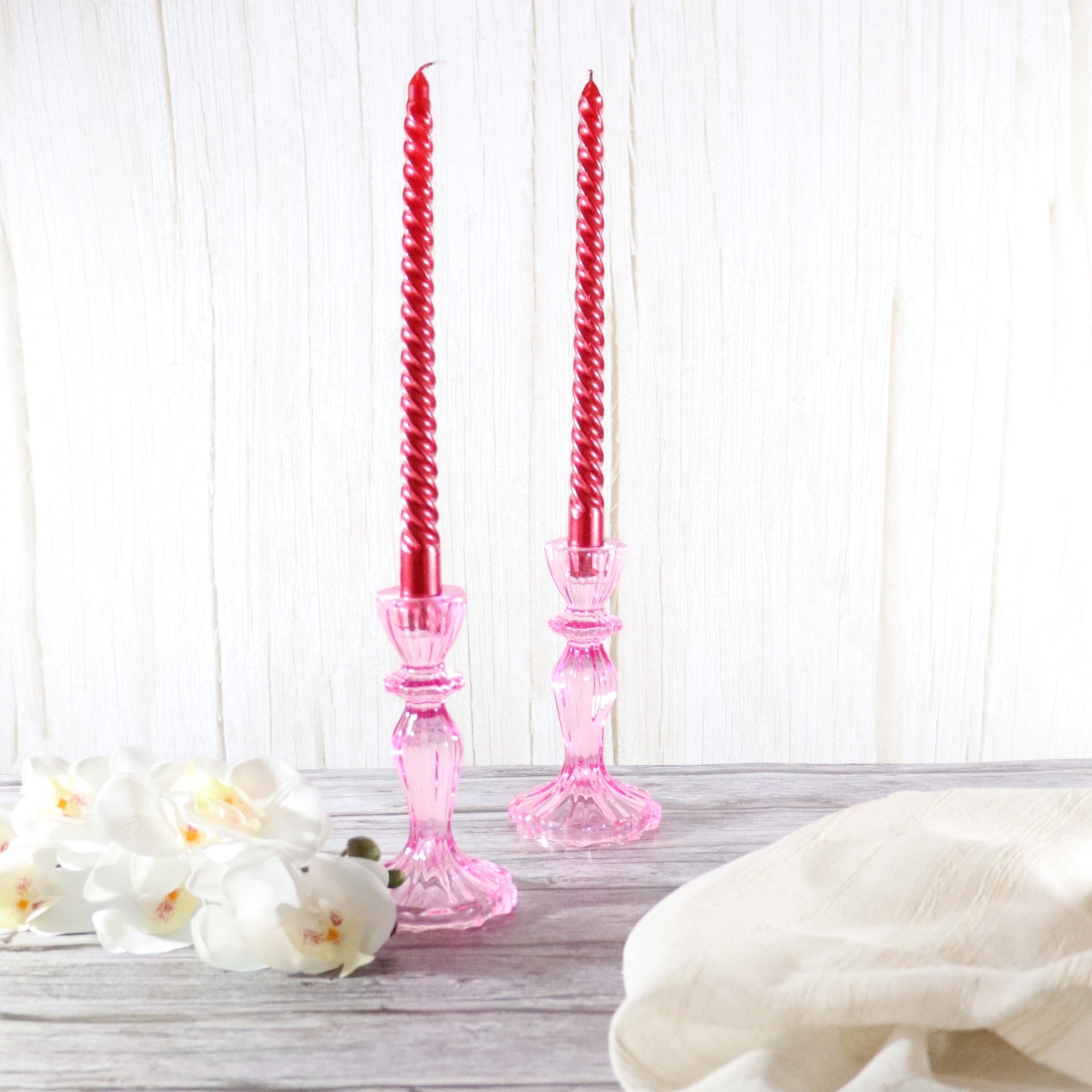 Red twisted candles in pink glass candlestick holders on a wooden surface with white flowers and a white cloth.