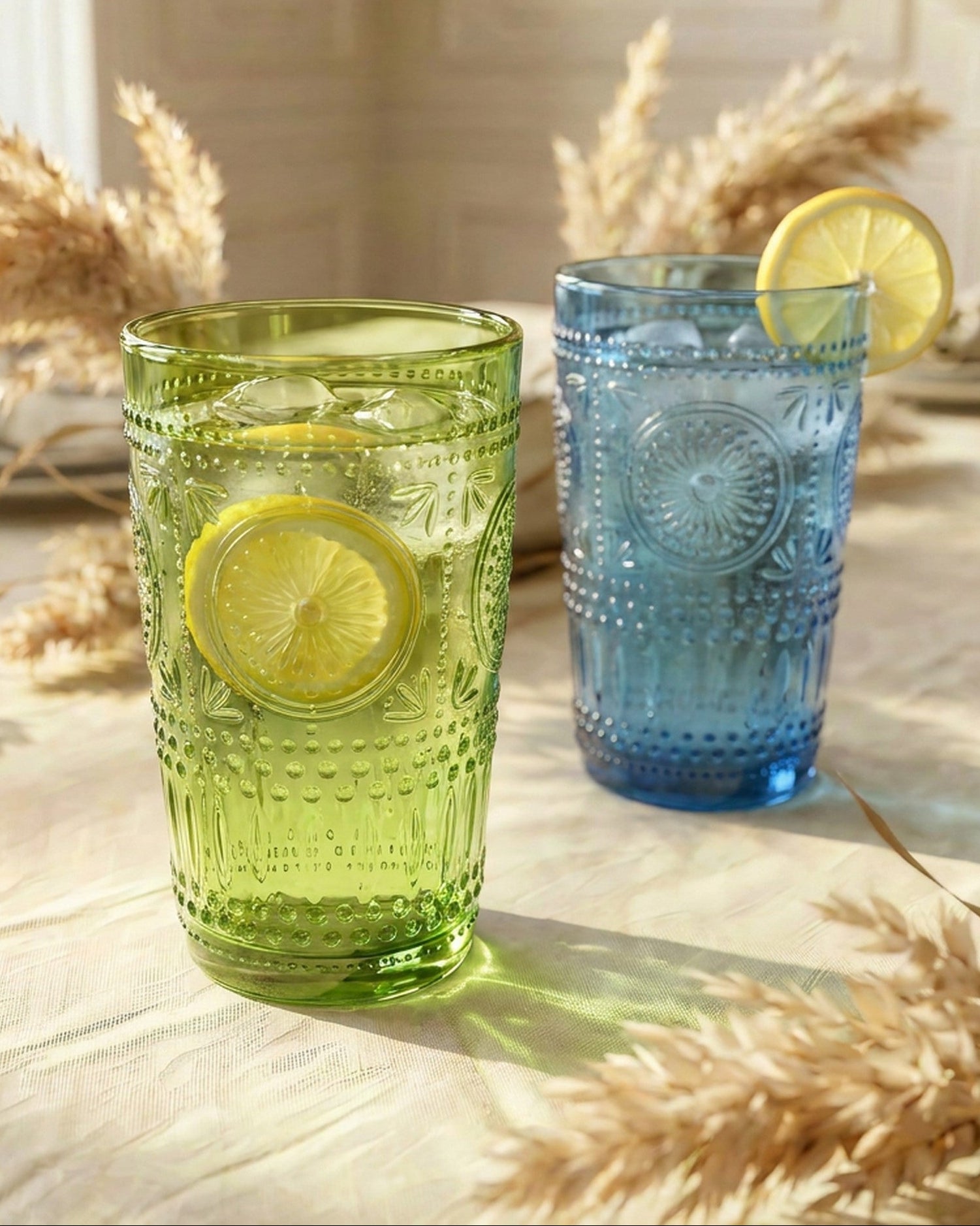 Two embossed glass tumblers on a table, one translucent light green and one translucent blue, both styled with lemon slices and surrounded by dried grasses in a softly lit interior setting.