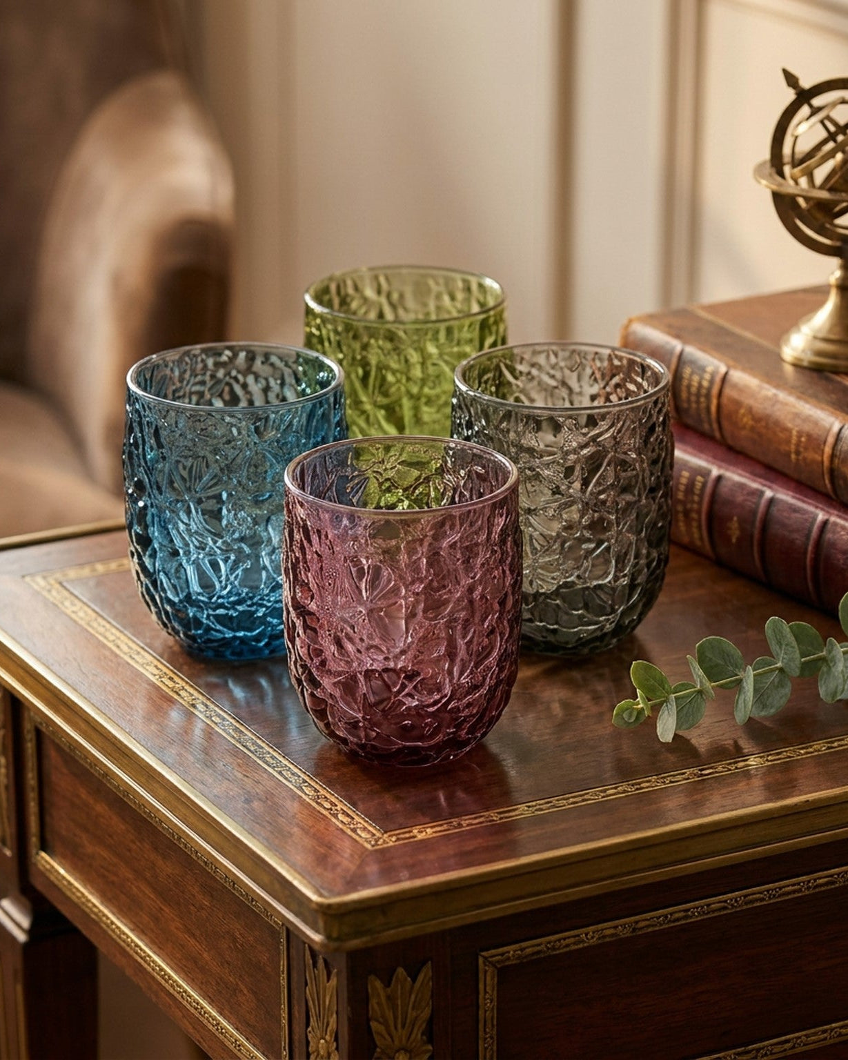 Four textured glass tumblers on a wooden table with books and a decorative sphere in the background.