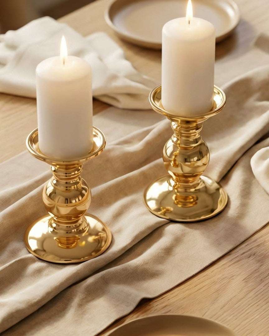 Two gold candle holders with lit candles on a wooden table with beige napkins and plates.