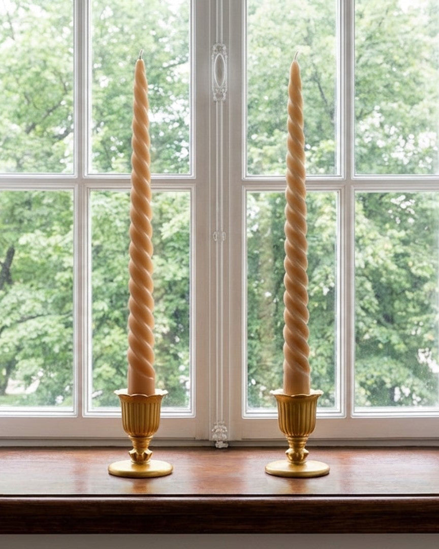 Two twisted candles in gold candle holders on a wooden surface with a window in the background showing greenery.