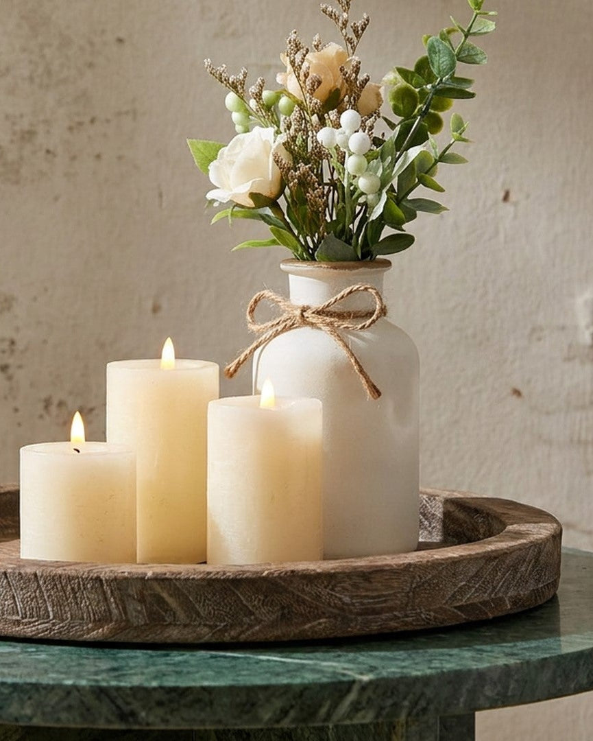 Decorative setting with candles and a vase of flowers on a round wooden tray against a textured wall.