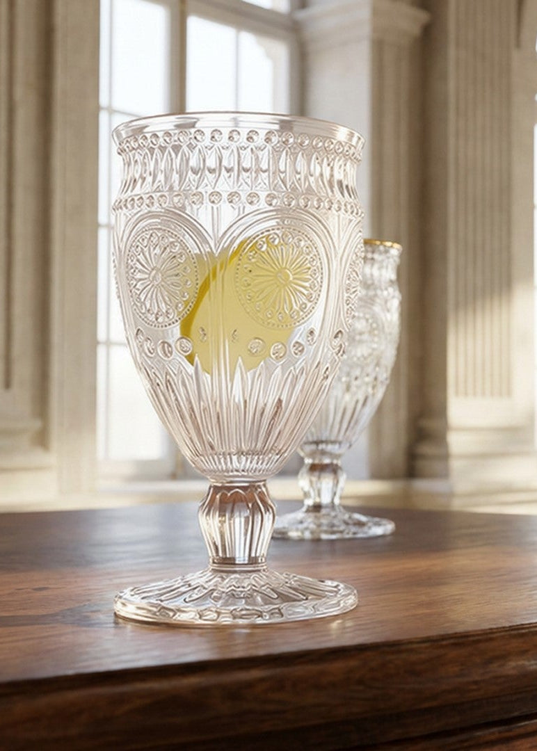 A clear cut-glass goblet with a yellow heart design on the front, displayed on a dark wooden table in a grand, sunlit classical room with arched windows.