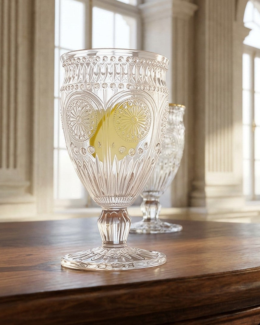 A clear cut-glass goblet with a heart pattern and a yellow center medallion, displayed on a dark wooden table in a grand, sunlit classical room with arched windows.