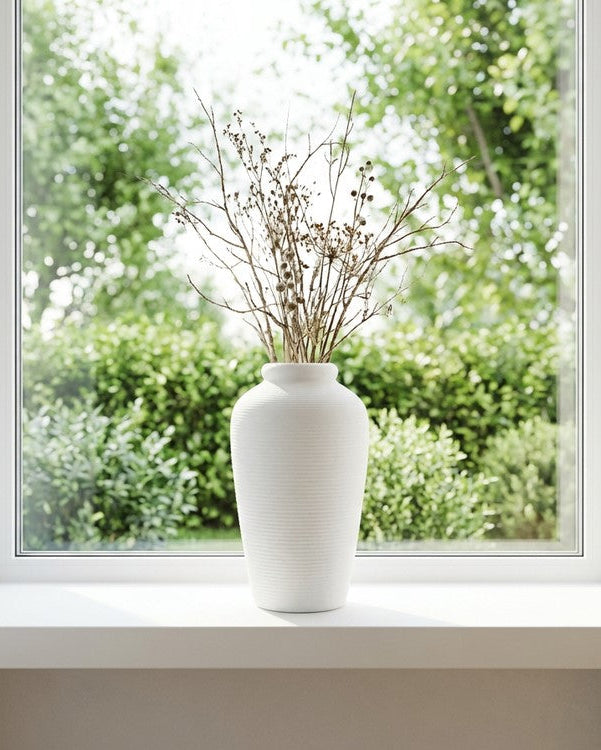 White vase with dried plants on a windowsill with greenery outside
