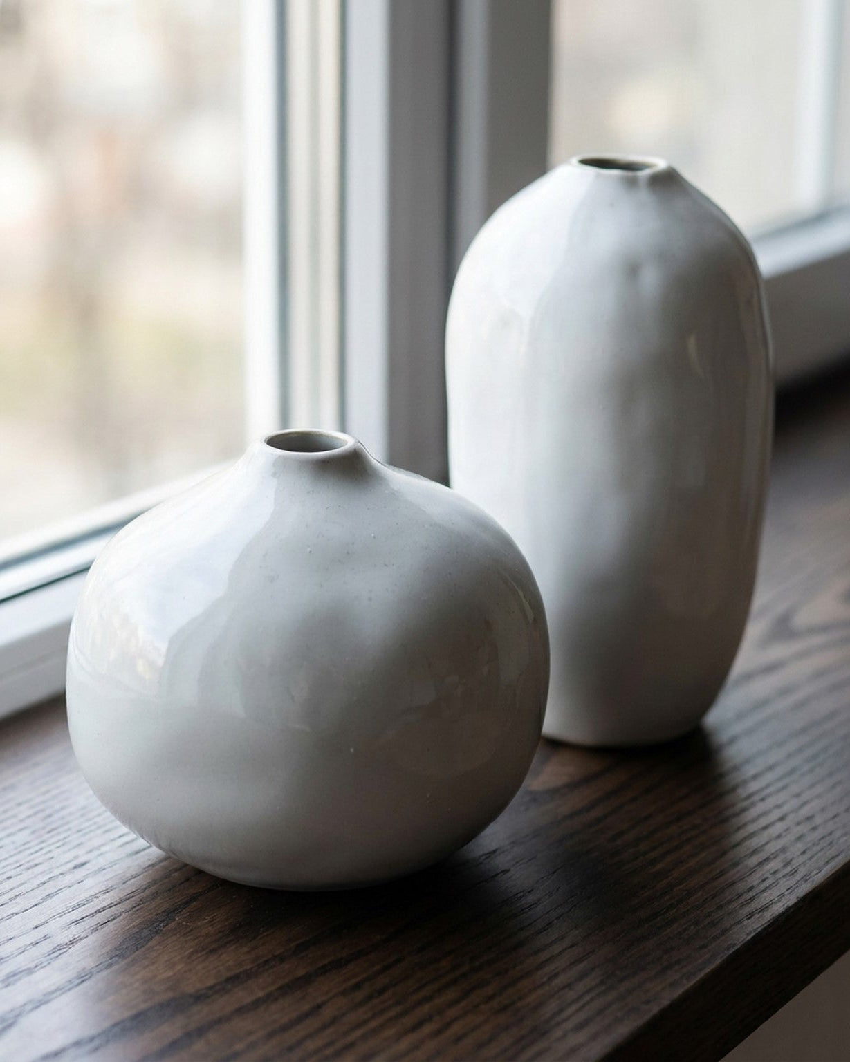 Two white ceramic vases on a wooden surface with a window in the background