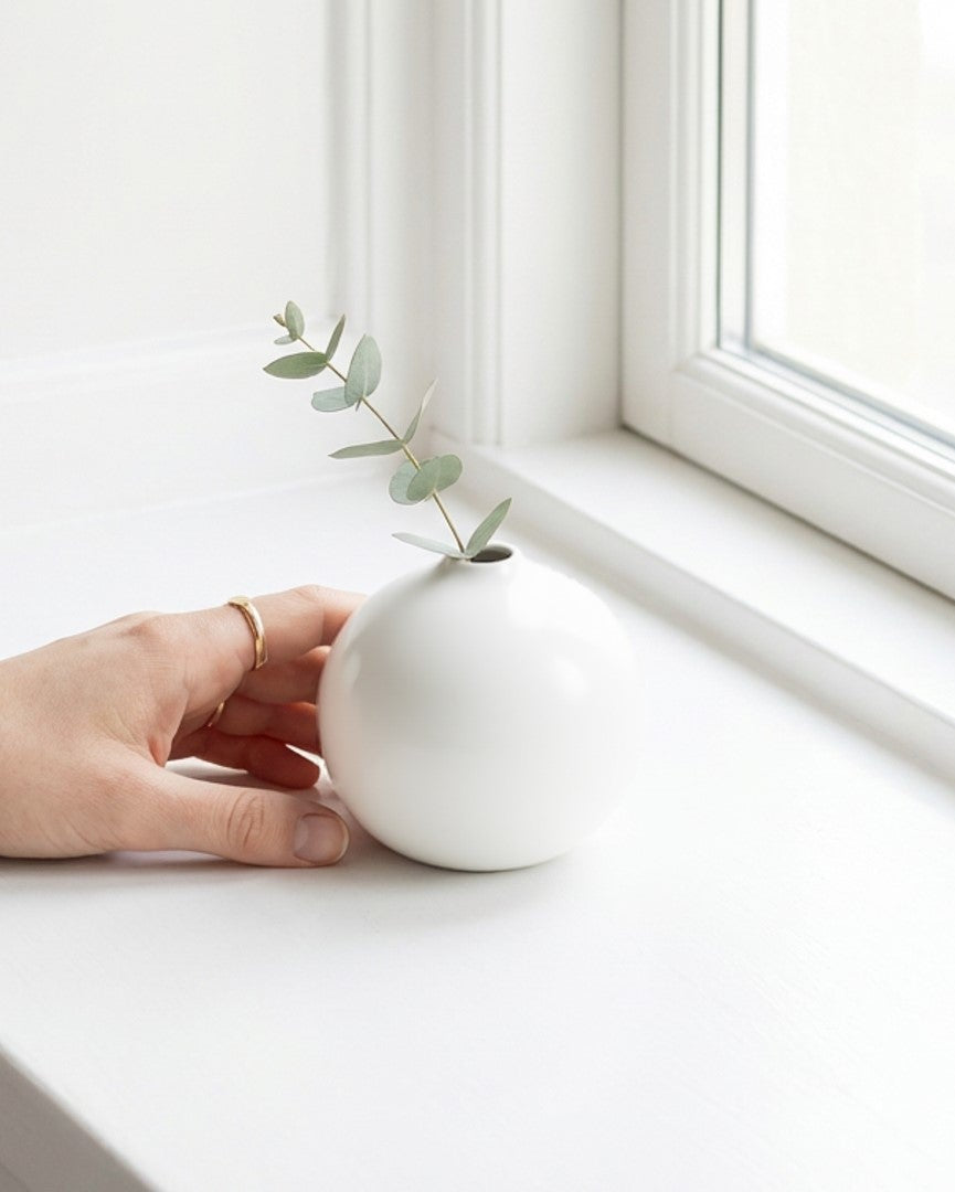 Person holding a small white vase with a plant on a windowsill.