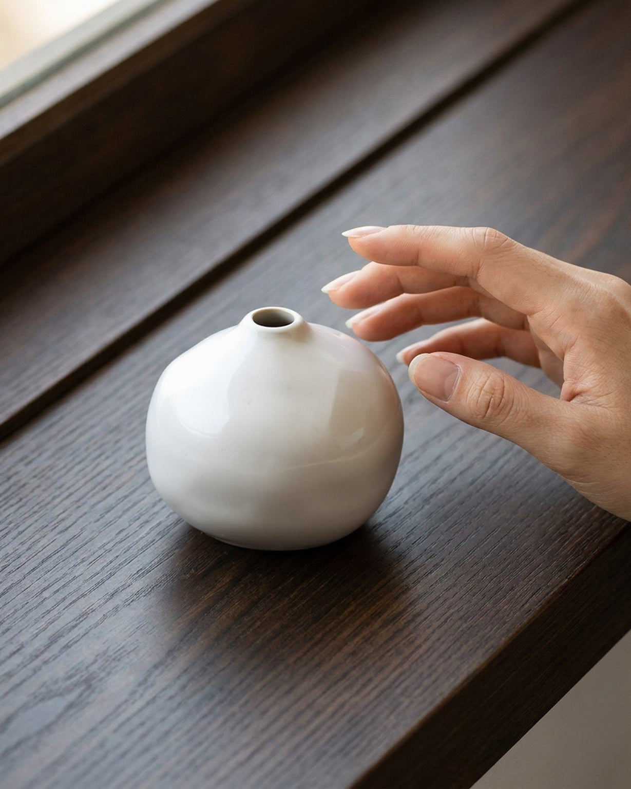 White ceramic vase on a wooden surface with a hand reaching towards it