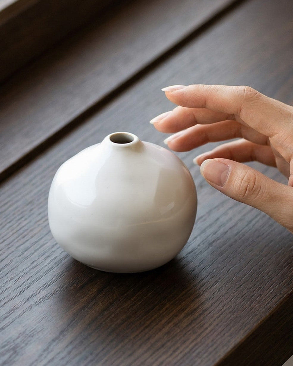 White ceramic vase on a wooden surface with a hand reaching towards it