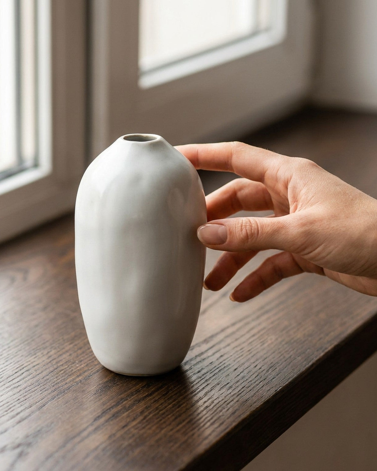 Hand touching a small white vase on a wooden surface with a window in the background