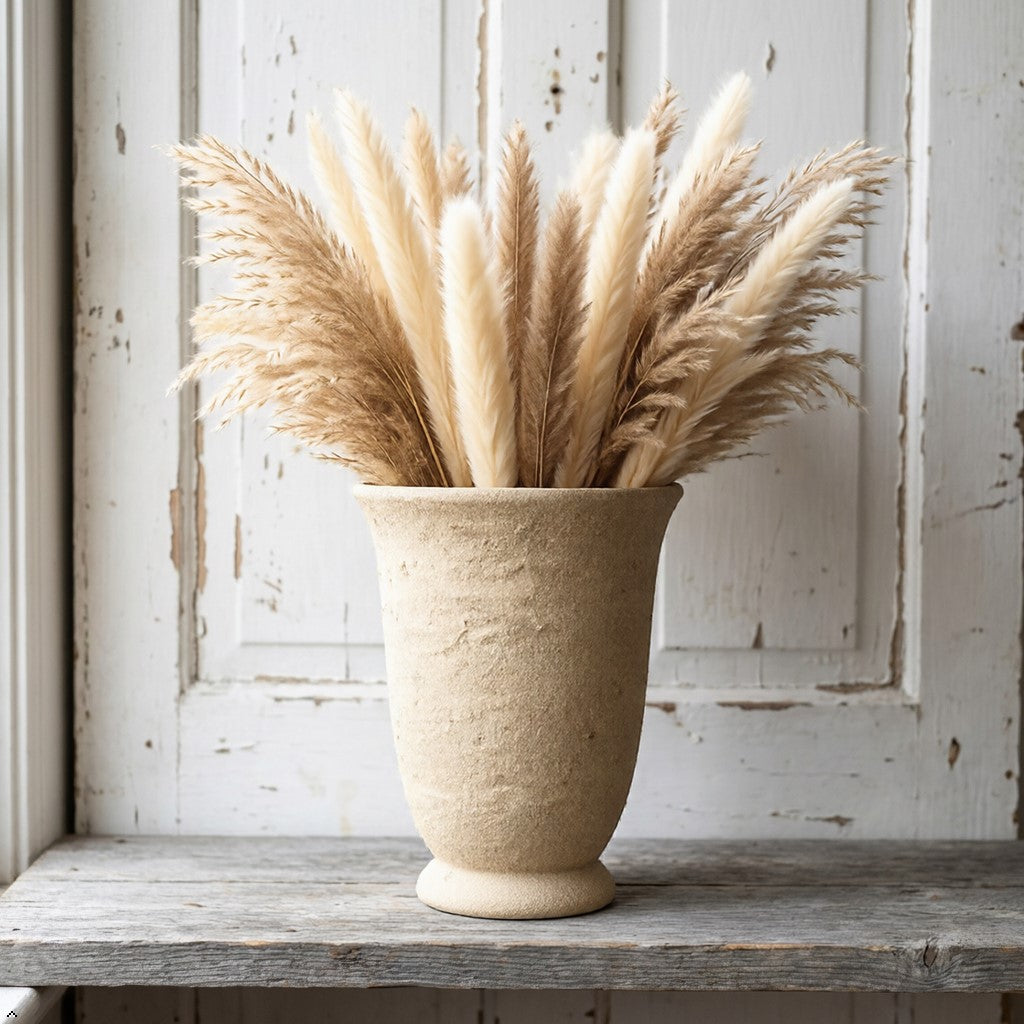Beige vase with dried pampas grass on a wooden surface against a white distressed wall.