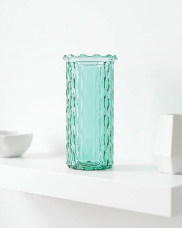 Turquoise glass vase on a white surface with a white bowl and geometric sculpture in the background.