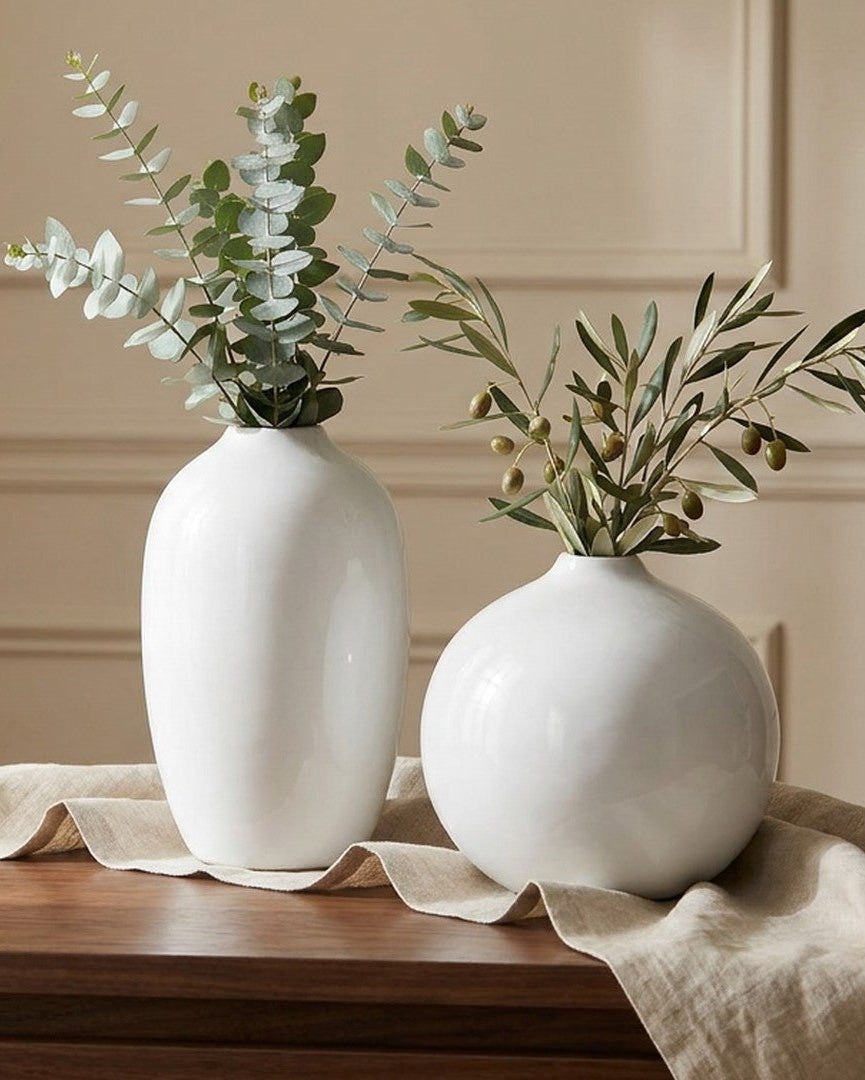 Two white vases with greenery on a wooden table against a beige wall.
