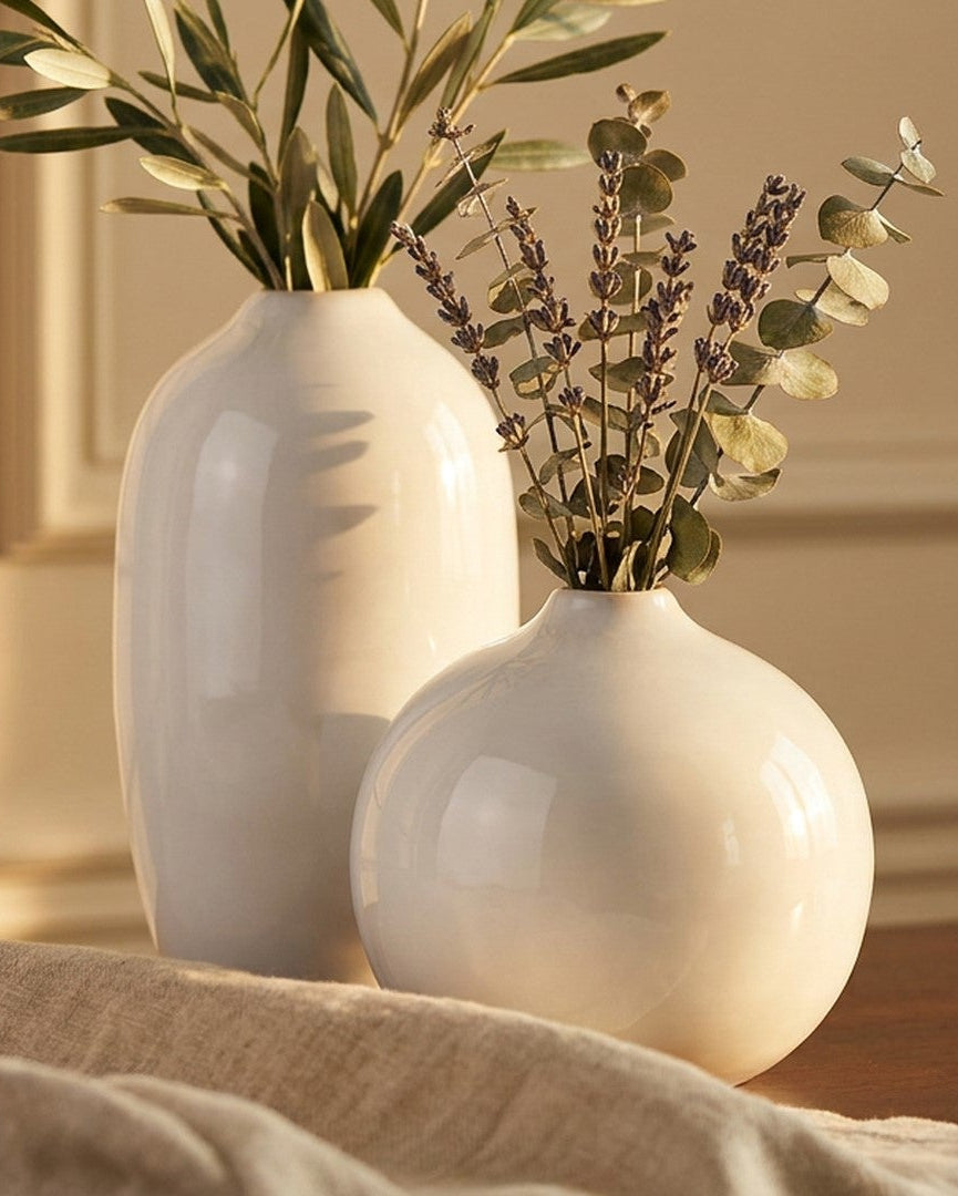 Two white vases with plants on a wooden surface against a beige wall.