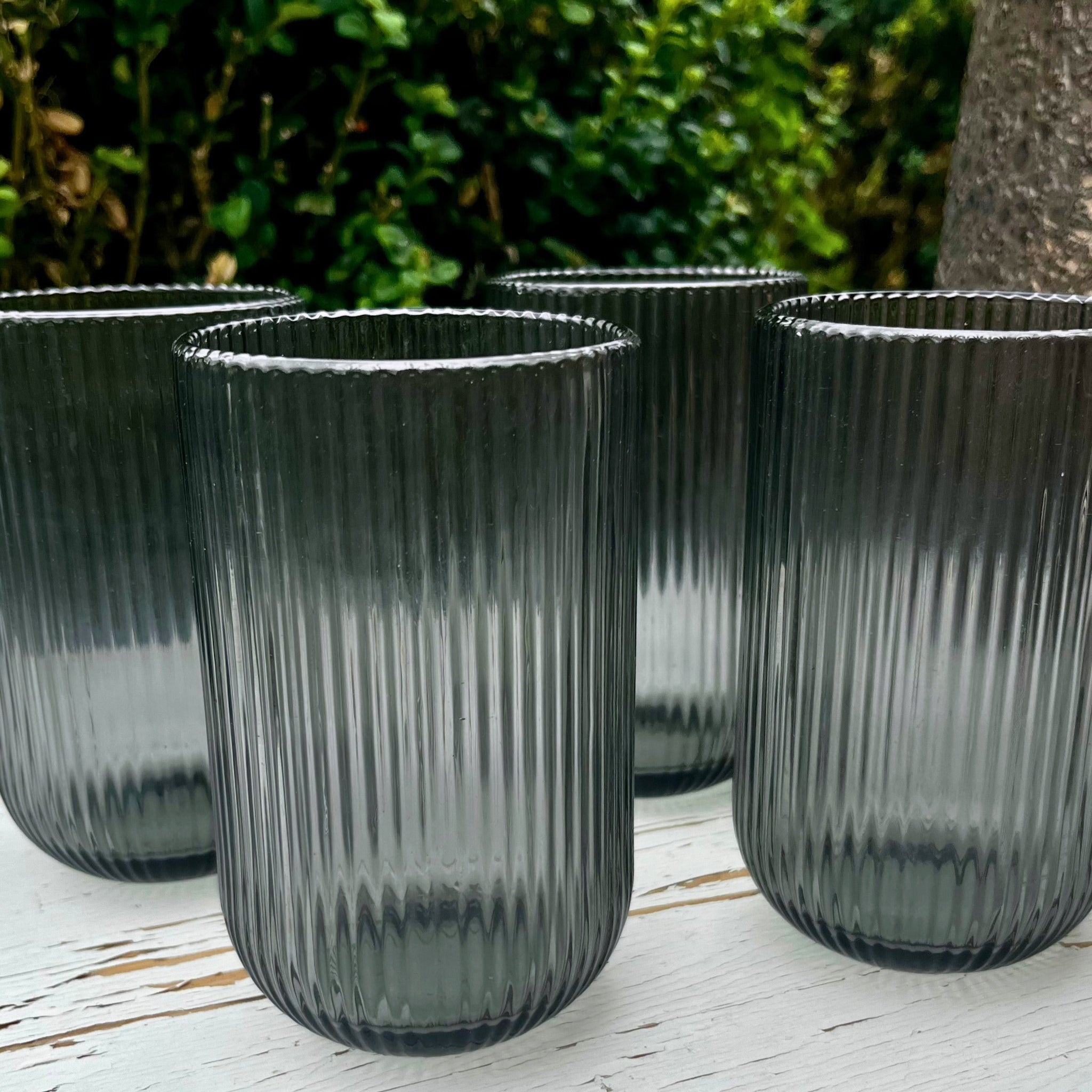 Set of four ribbed dark gray glass tumblers on a wooden surface with a blurred green outdoor background.