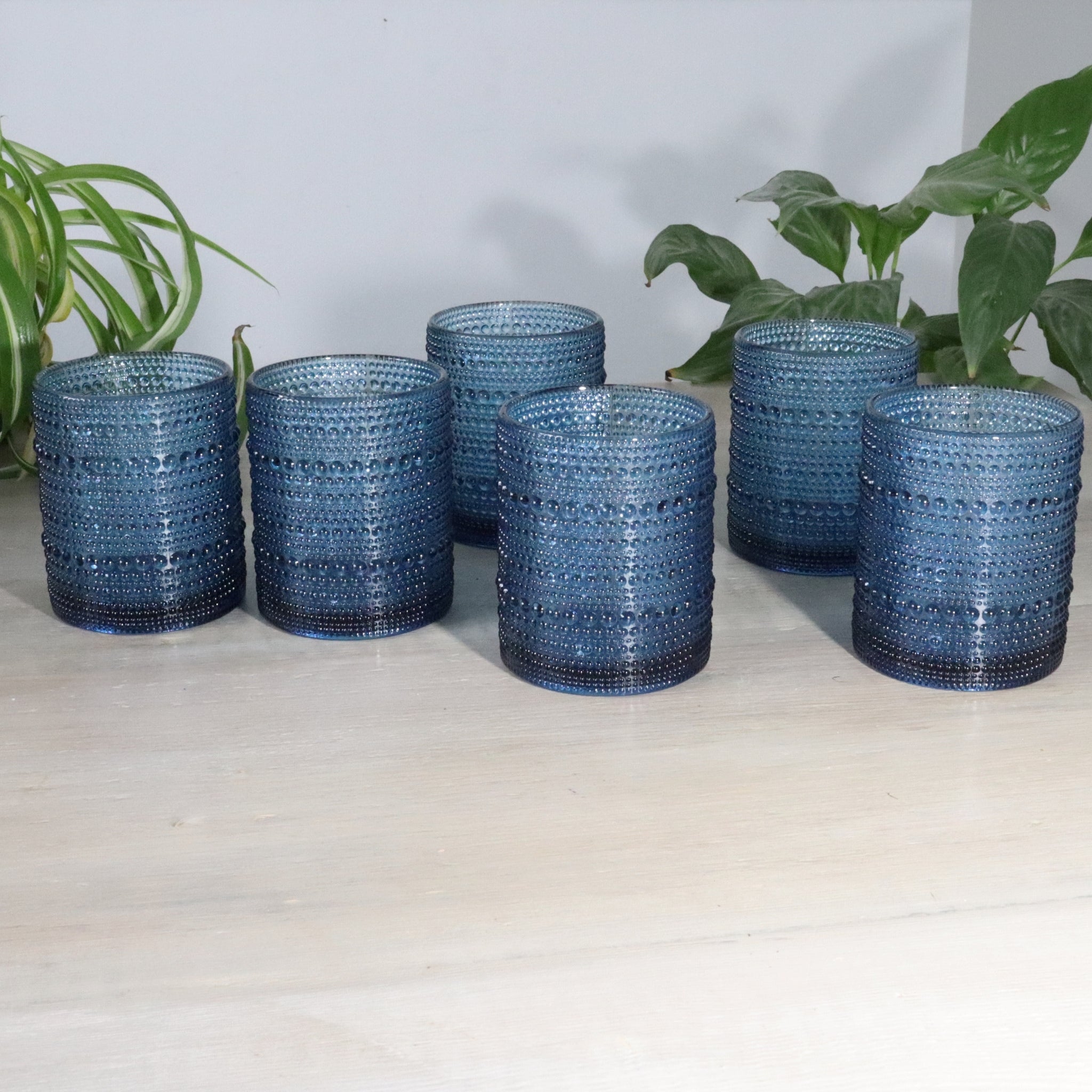Set of blue hobnail glass tumblers on a light surface with plants in the background