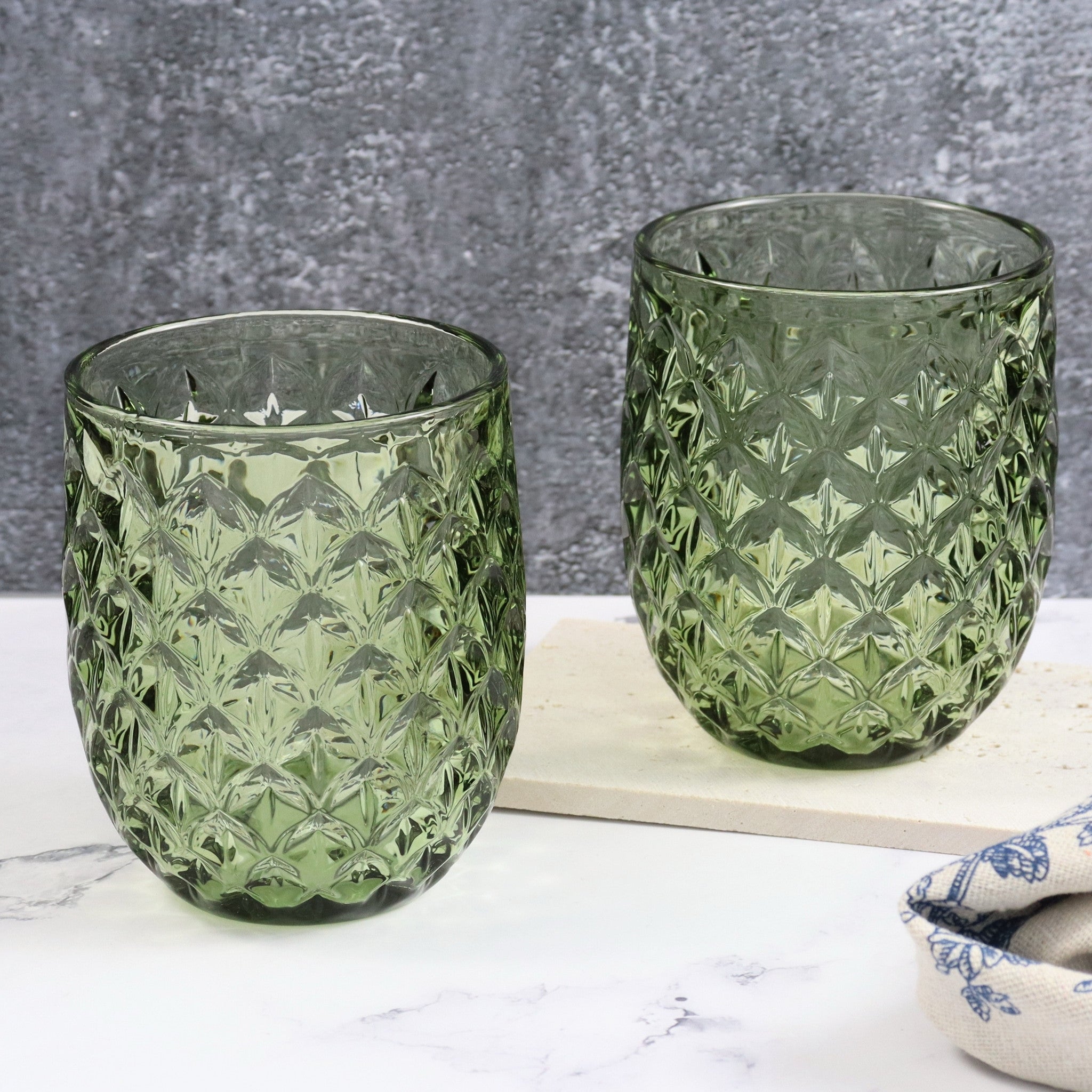Two green patterned glass tumblers on a marble surface with a gray textured background.
