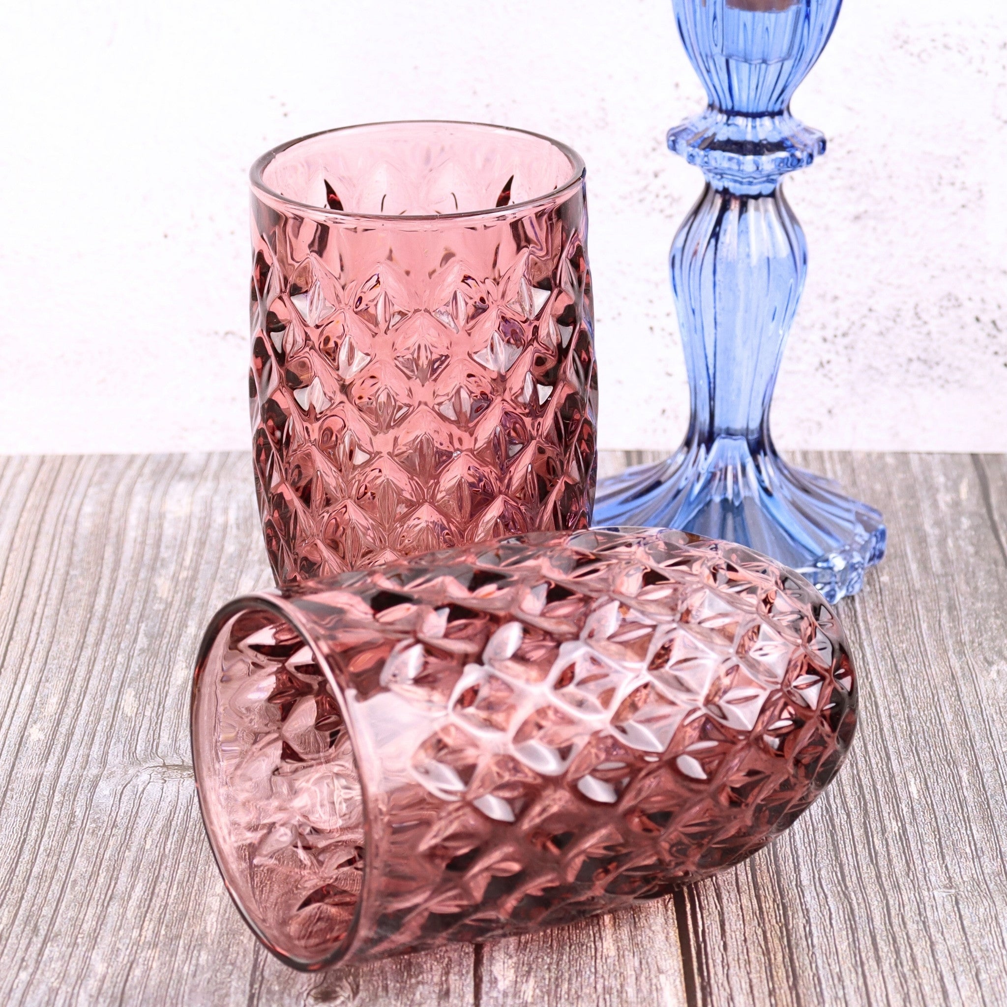 Two pink textured glass tumblers and a blue glass candle holder on a wooden surface.