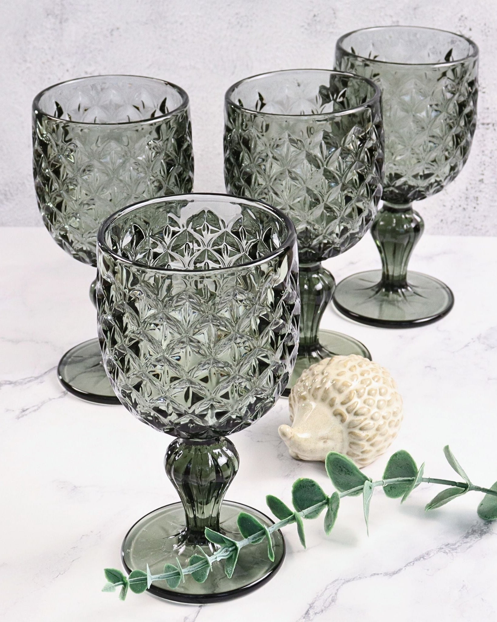 Set of four grey glass goblets on a marble surface with a small decorative shell and eucalyptus leaf.
