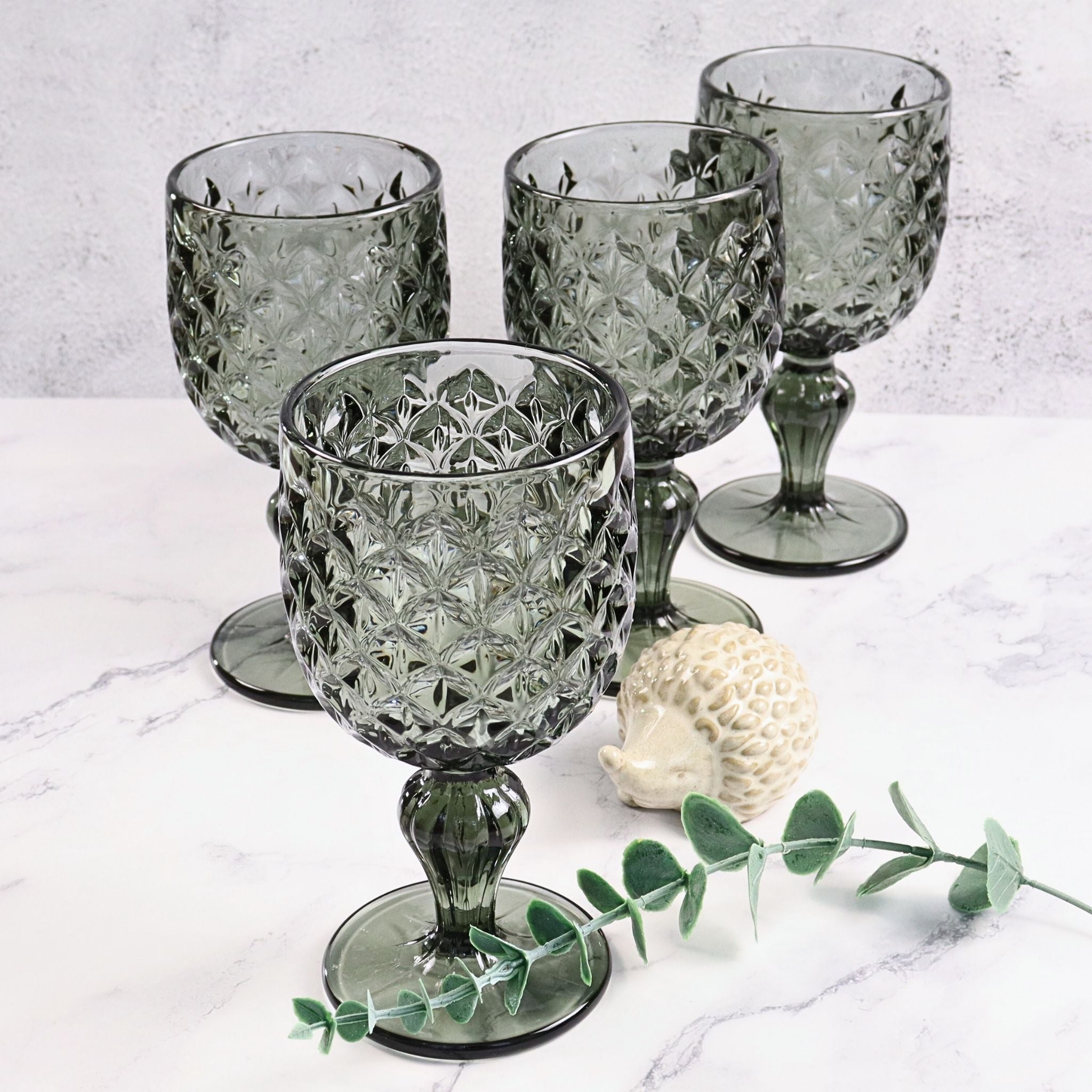 Set of four grey glass goblets on a marble surface with a small decorative shell and eucalyptus leaf.