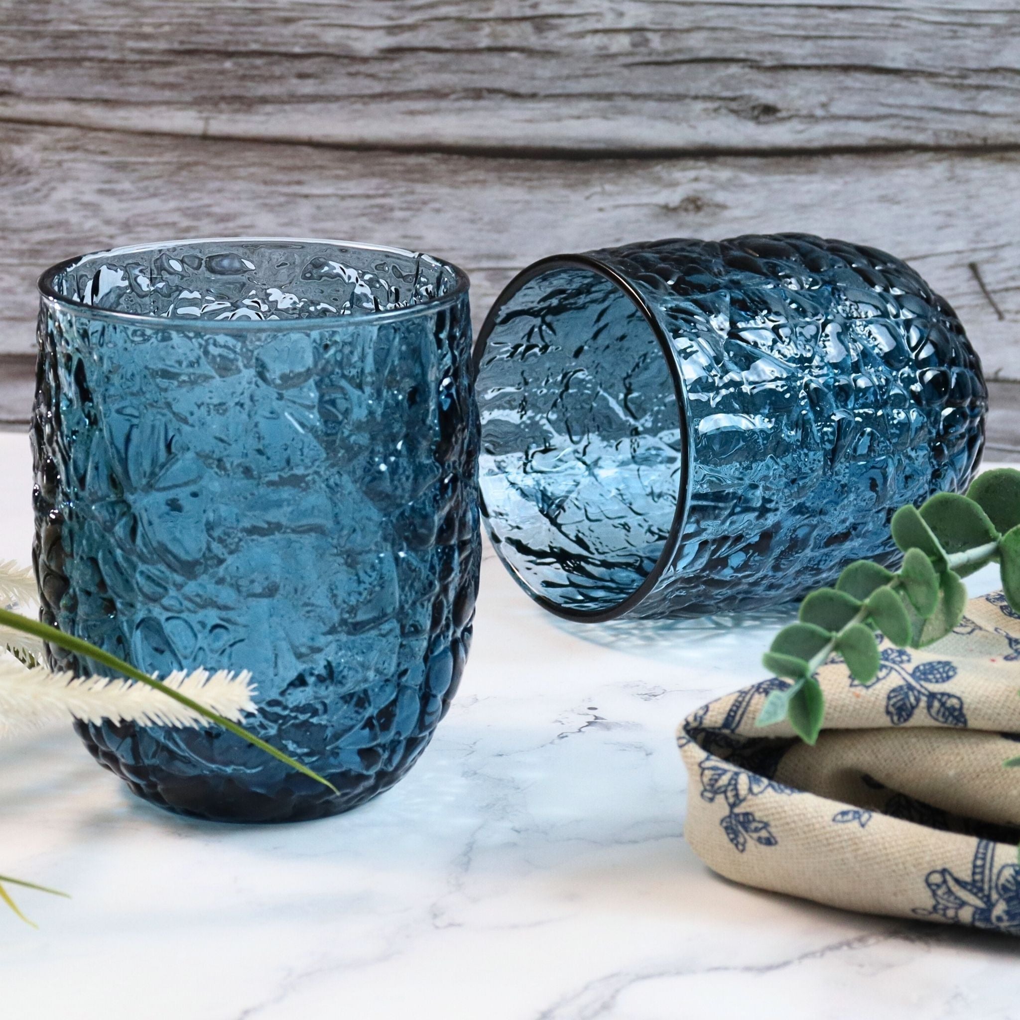 Two blue textured glass tumblers on a marble surface with a wooden background.
