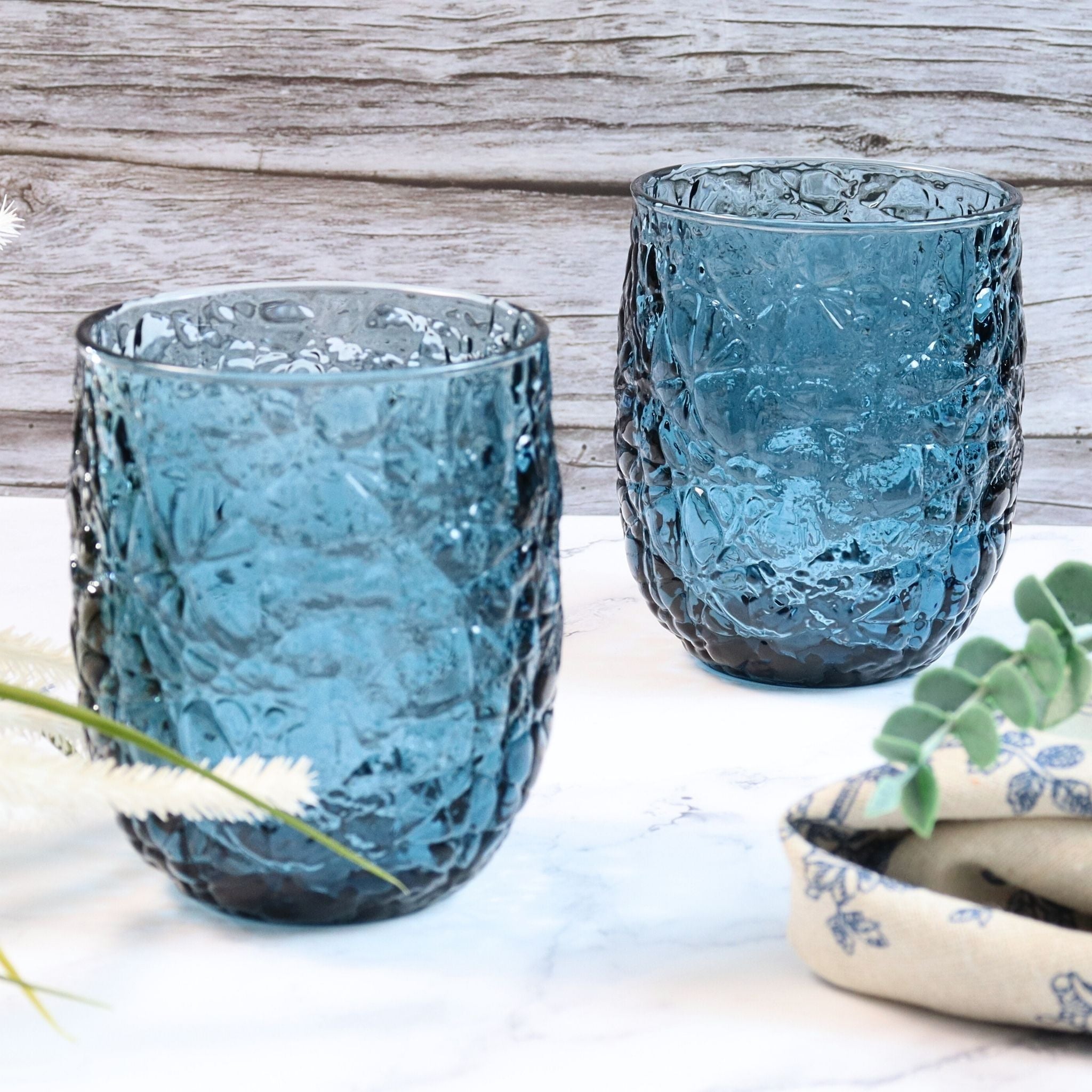 Two textured blue glass tumblers on a light wooden surface with a plant in the background.