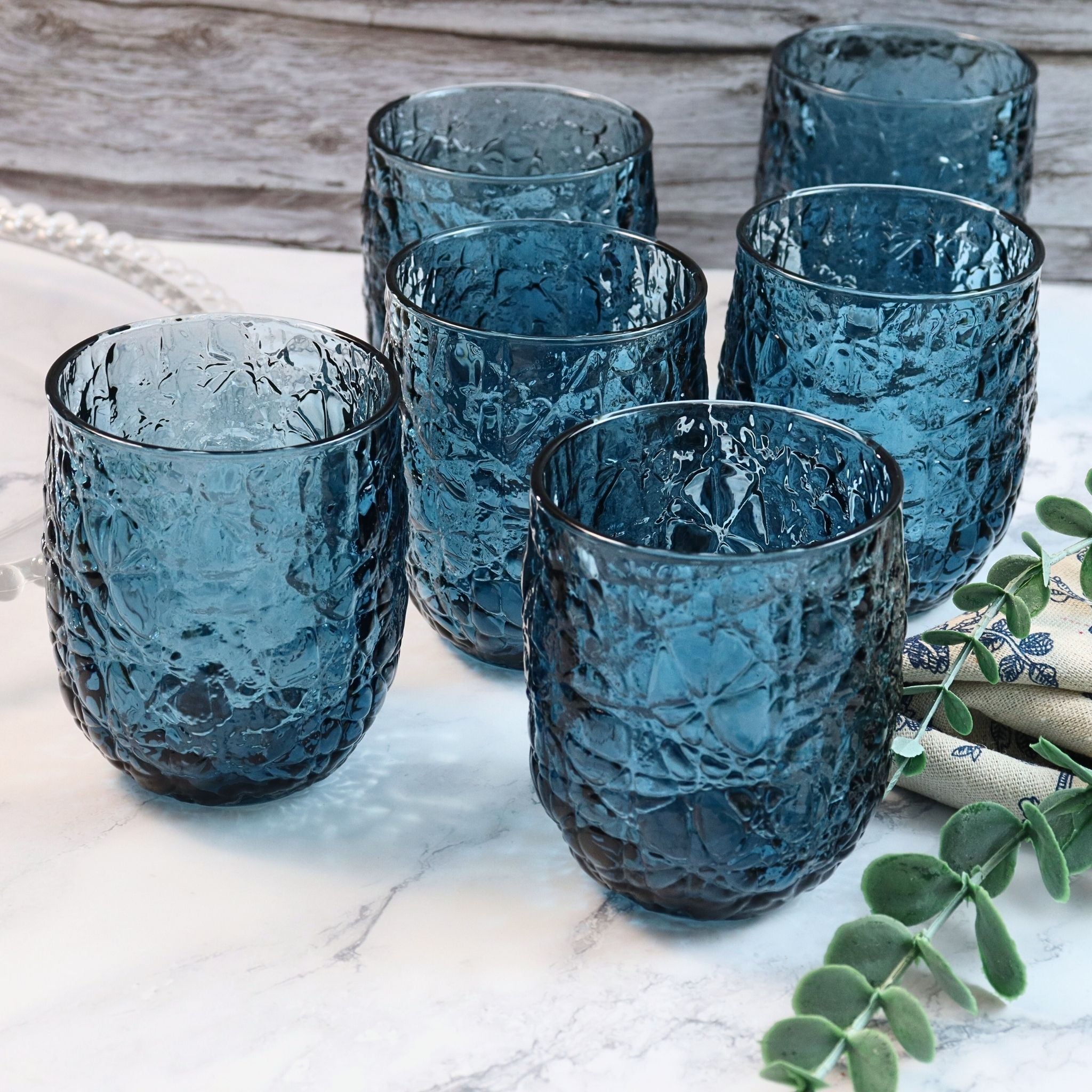 Set of six blue textured glass tumblers on a marble surface with a wooden background.