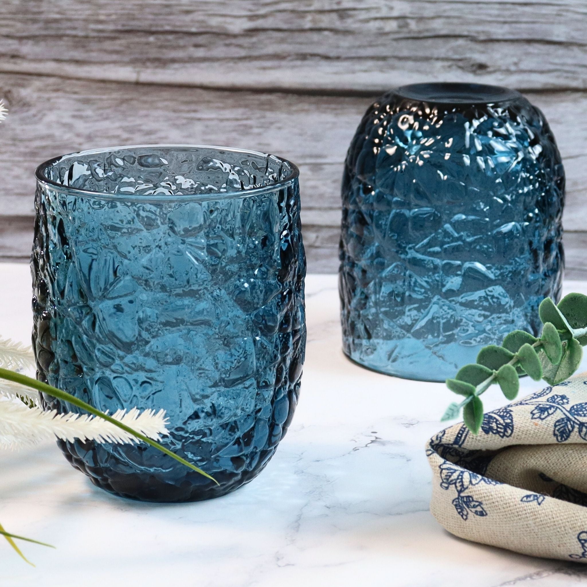 Two textured blue glass containers on a marble surface with a wooden background