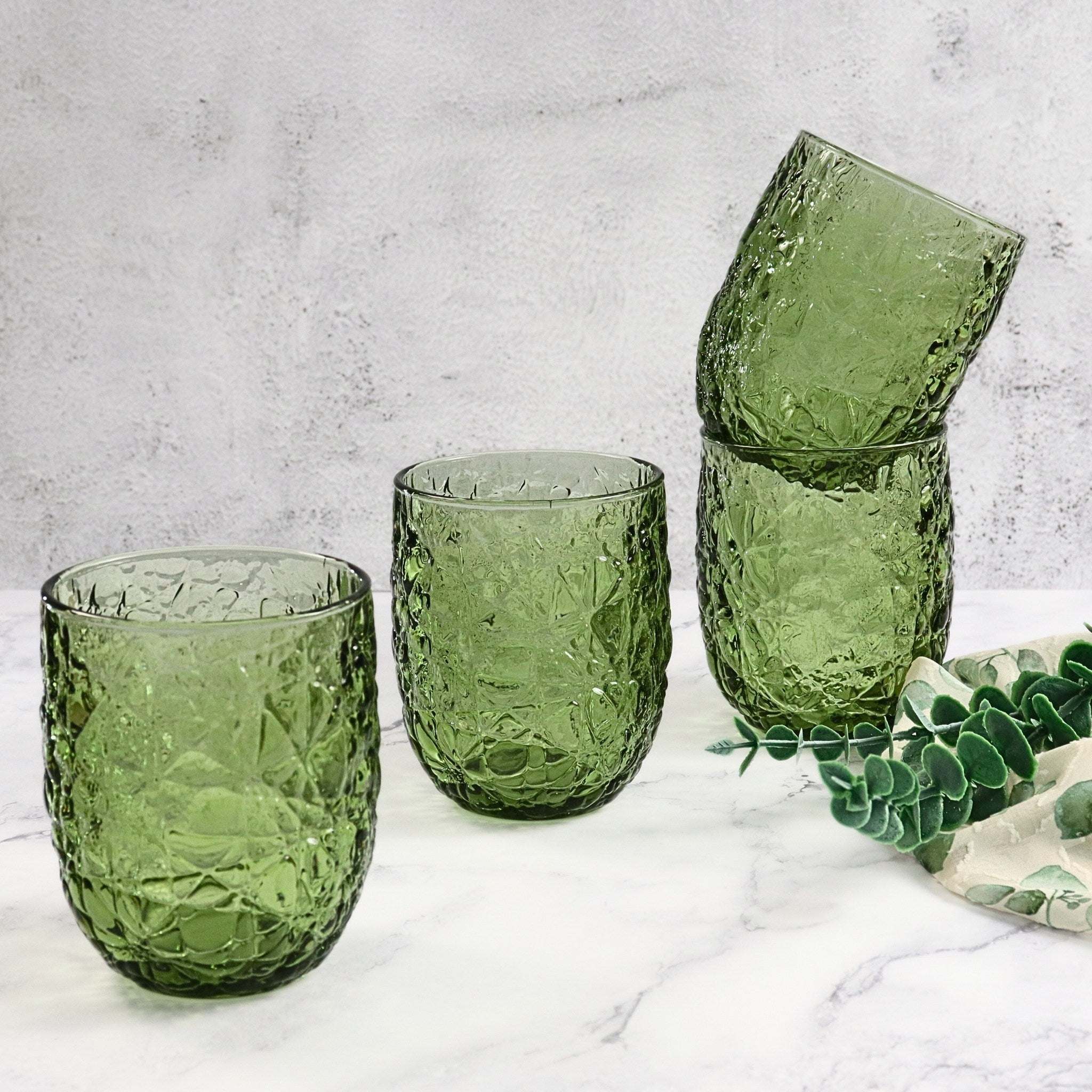 Set of green textured glass tumblers on a light gray surface with a plant in the background.