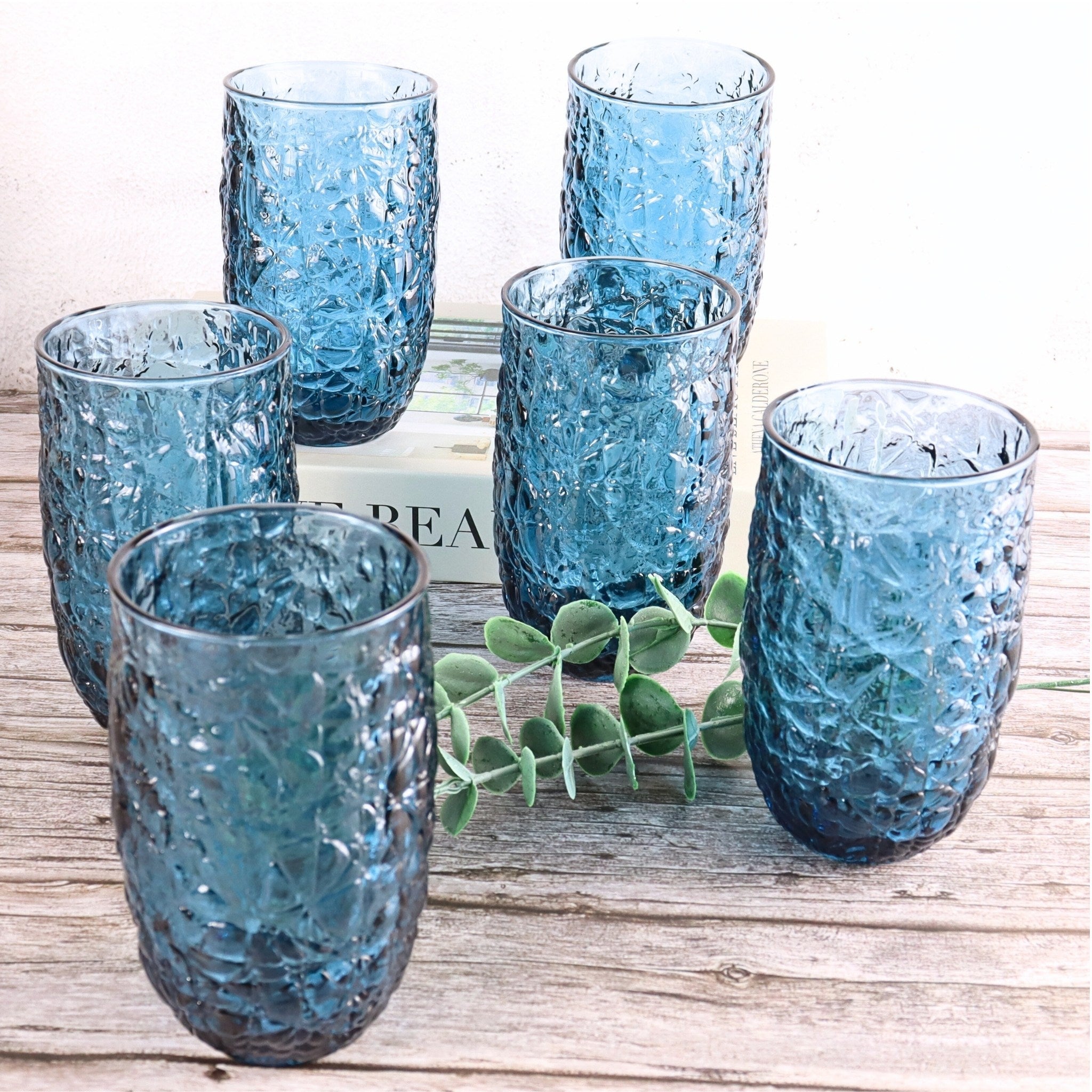 Set of six blue textured glass tumblers on a wooden surface with a plant in the background.
